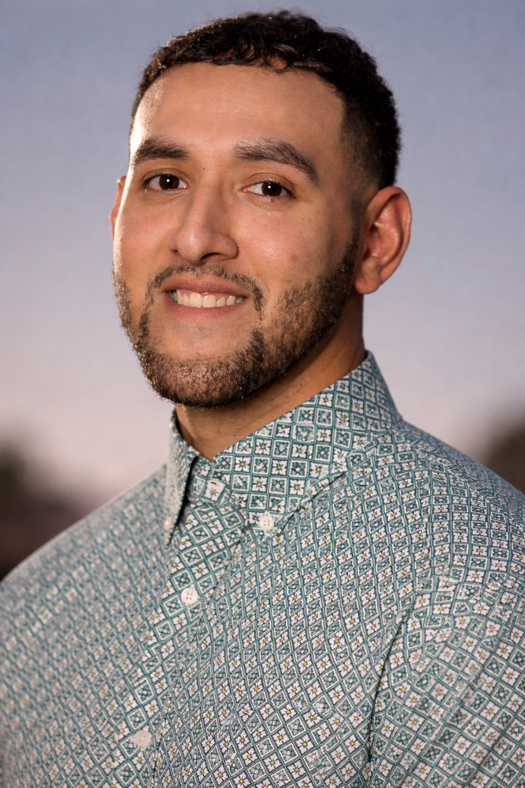 Close-up of a man with short curly hair and a beard, smiling slightly, wearing a patterned button-down shirt against a blurry outdoor background.