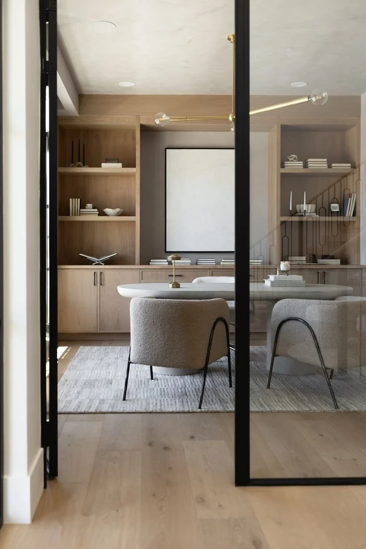 Modern dining room with a round white table, two gray upholstered chairs, built-in wooden shelves with books and decorative items, a large framed whiteboard or art piece on the wall, and a glass partition with black framing.