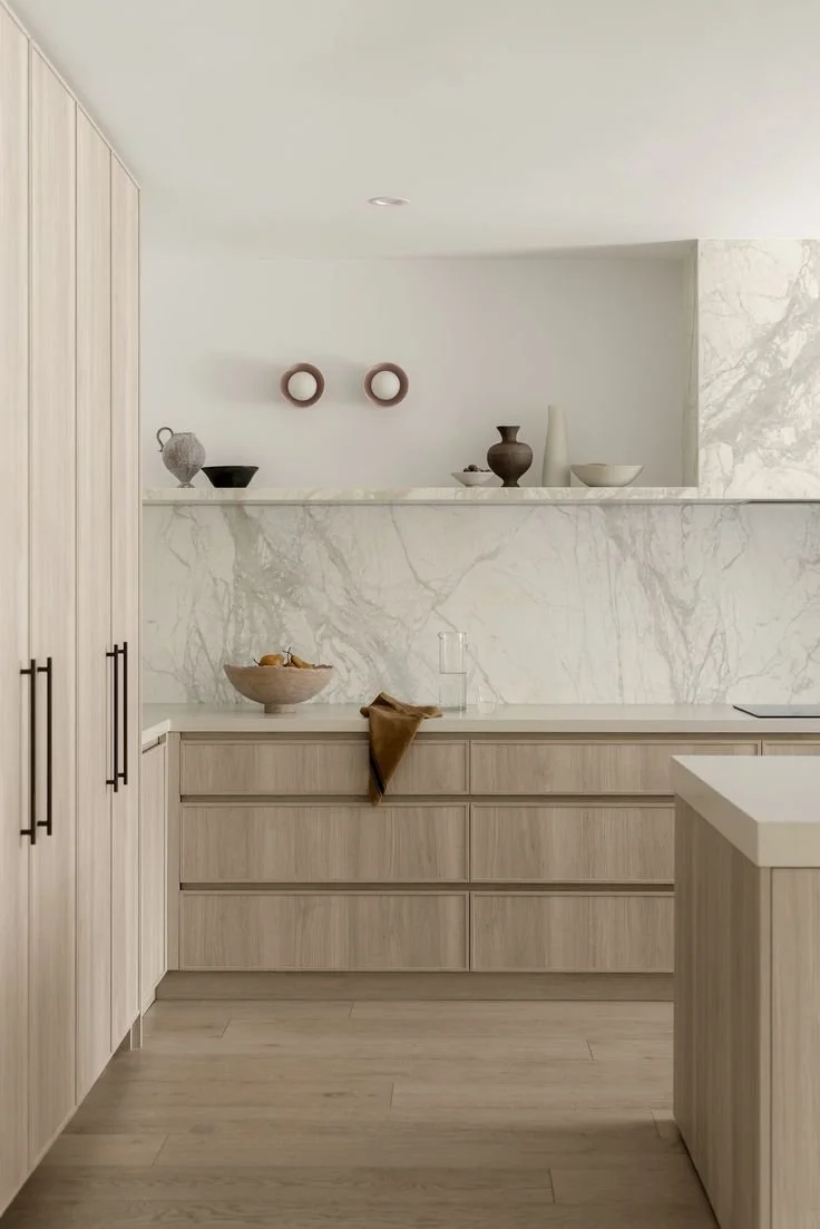 Minimalist kitchen with light wood cabinets, marble backsplash, and decorative pottery on the counter and a shelf.