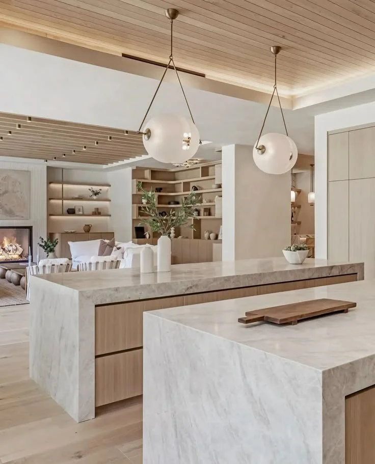 Modern, minimalist kitchen with marble countertops, wooden accents, and hanging globe pendant lights. The open living space features built-in shelving, a fireplace, and a seating area with pillows.