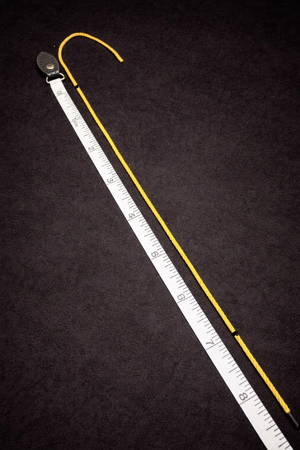 A yellow string with black marker markings and a black surface, positioned next to a white ruler marked in inches for size reference.