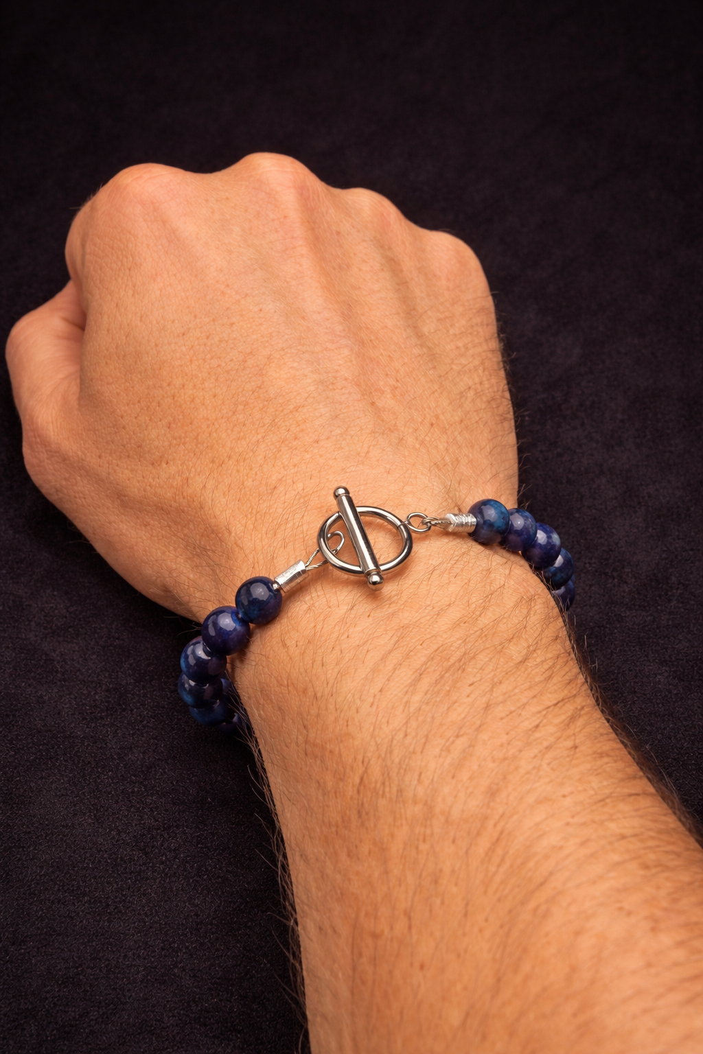 A person's wrist with a blue beaded bracelet featuring a toggle clasp. The background is black.