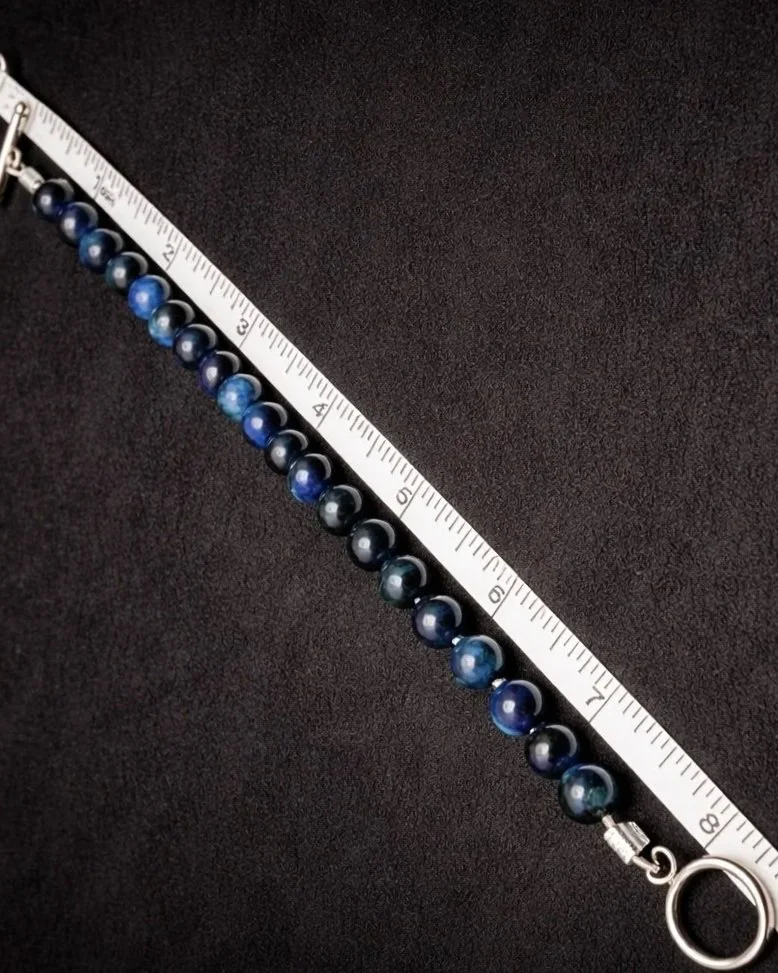 A necklace made of blue and black marbled beads laid next to a measuring tape on a black surface.