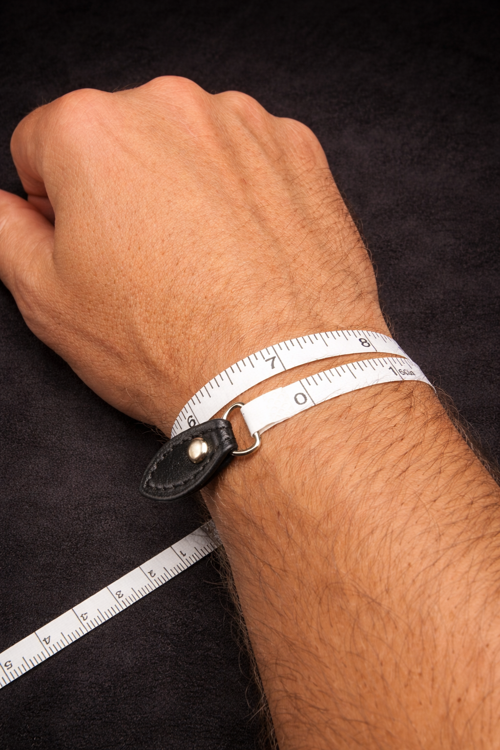 A person's hand and wrist on a black background, with a white measuring tape wrapped around the wrist showing approximately 7 inches.