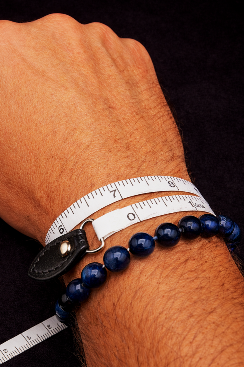 Close-up of a human wrist with a black leather watch strap, a white measuring tape, and a dark blue bead bracelet on a black background.
