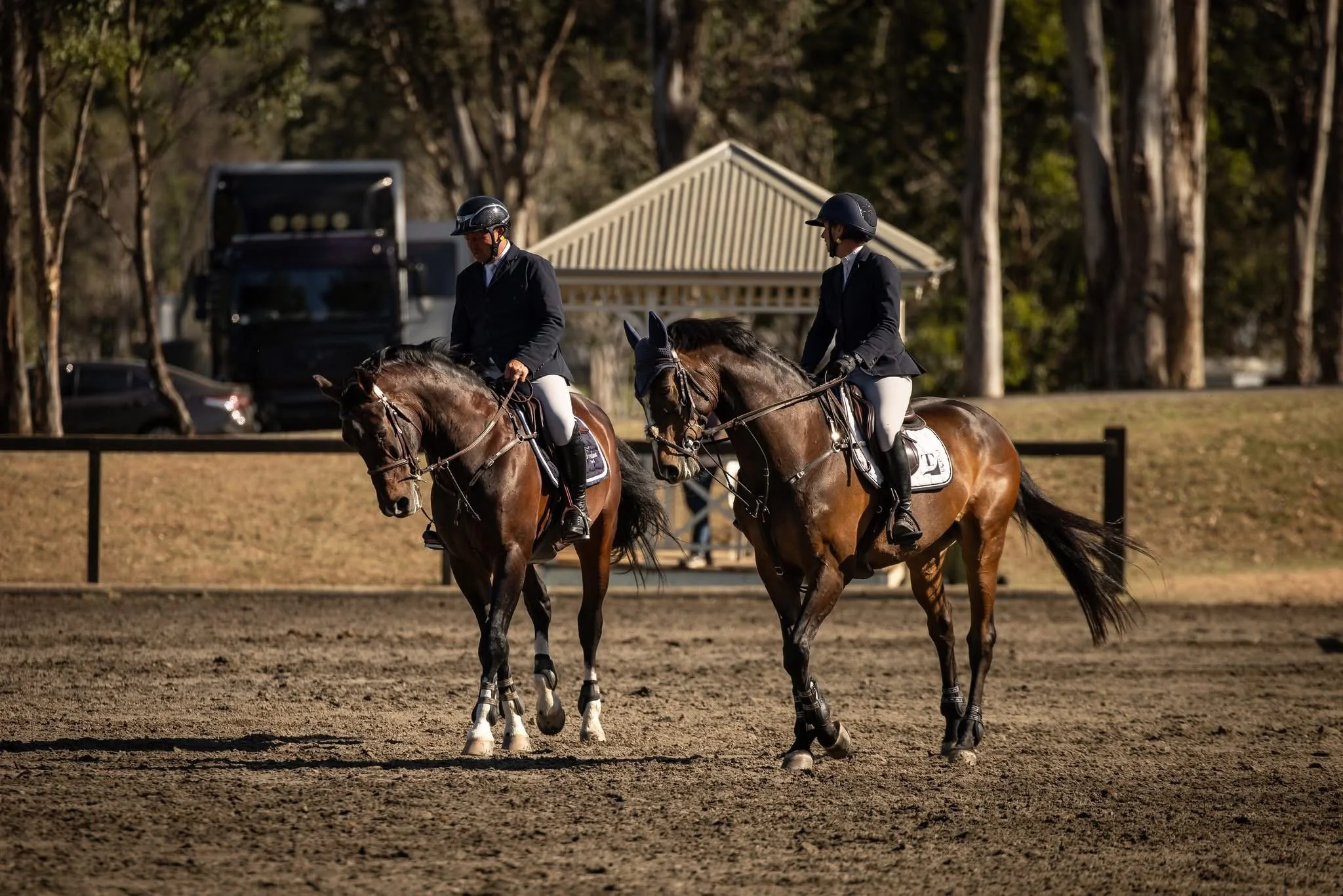 Two equestrians in formal riding attire on horseback on a dirt arena, with trees and a pavilion in the background.