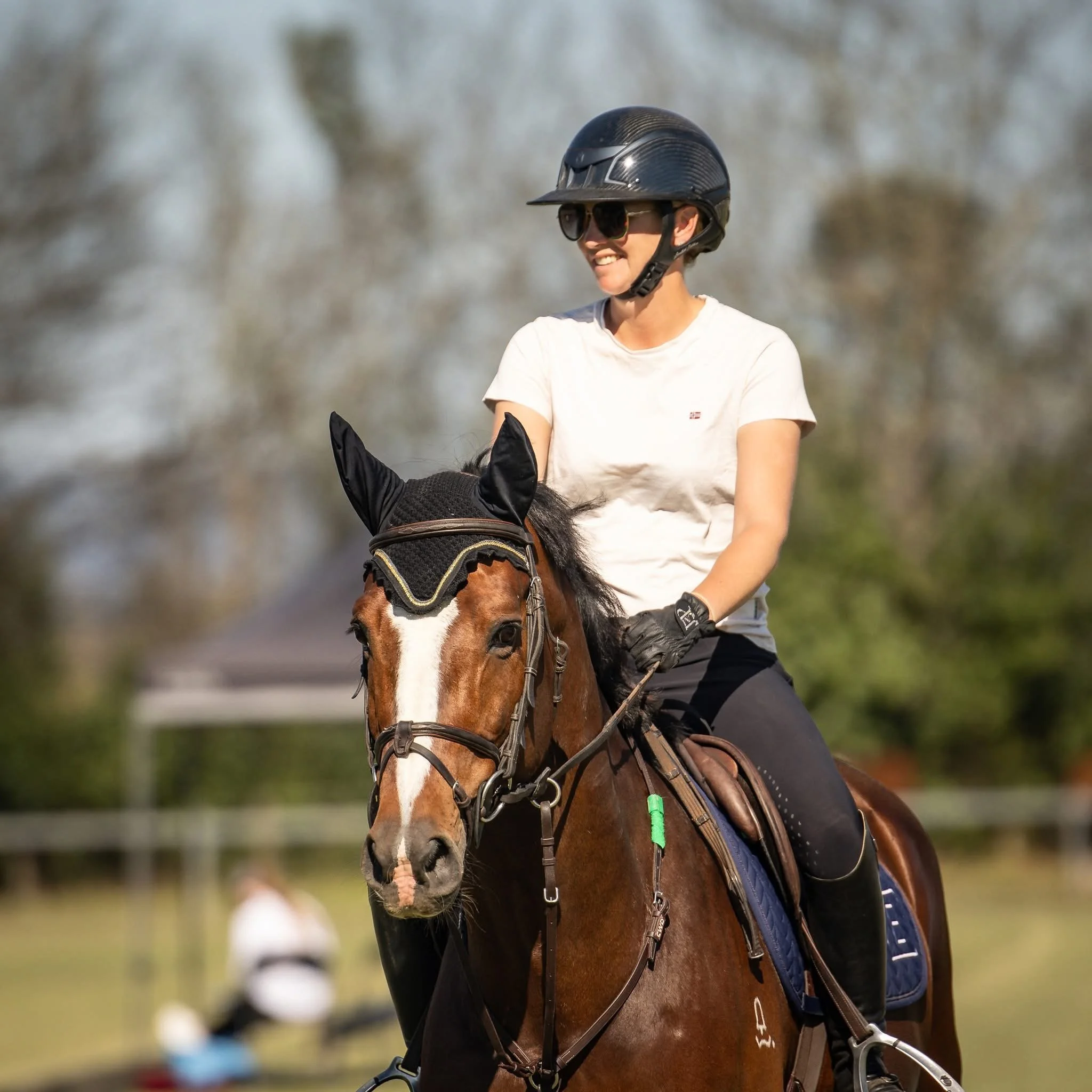 Woman in white t-shirt, black riding pants, helmet, and sunglasses riding a brown horse with a black mane in an outdoor setting with trees.