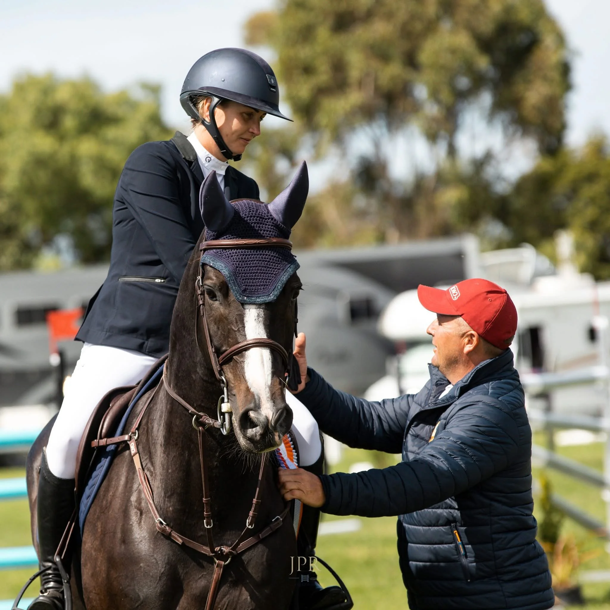 A female equestrian rider wearing a black helmet and riding jacket seated on a dark brown horse with a white blaze. The horse is wearing a mesh ear bonnet. A man in a red baseball cap and navy jacket is standing beside the horse, speaking to and gesturing toward the rider.