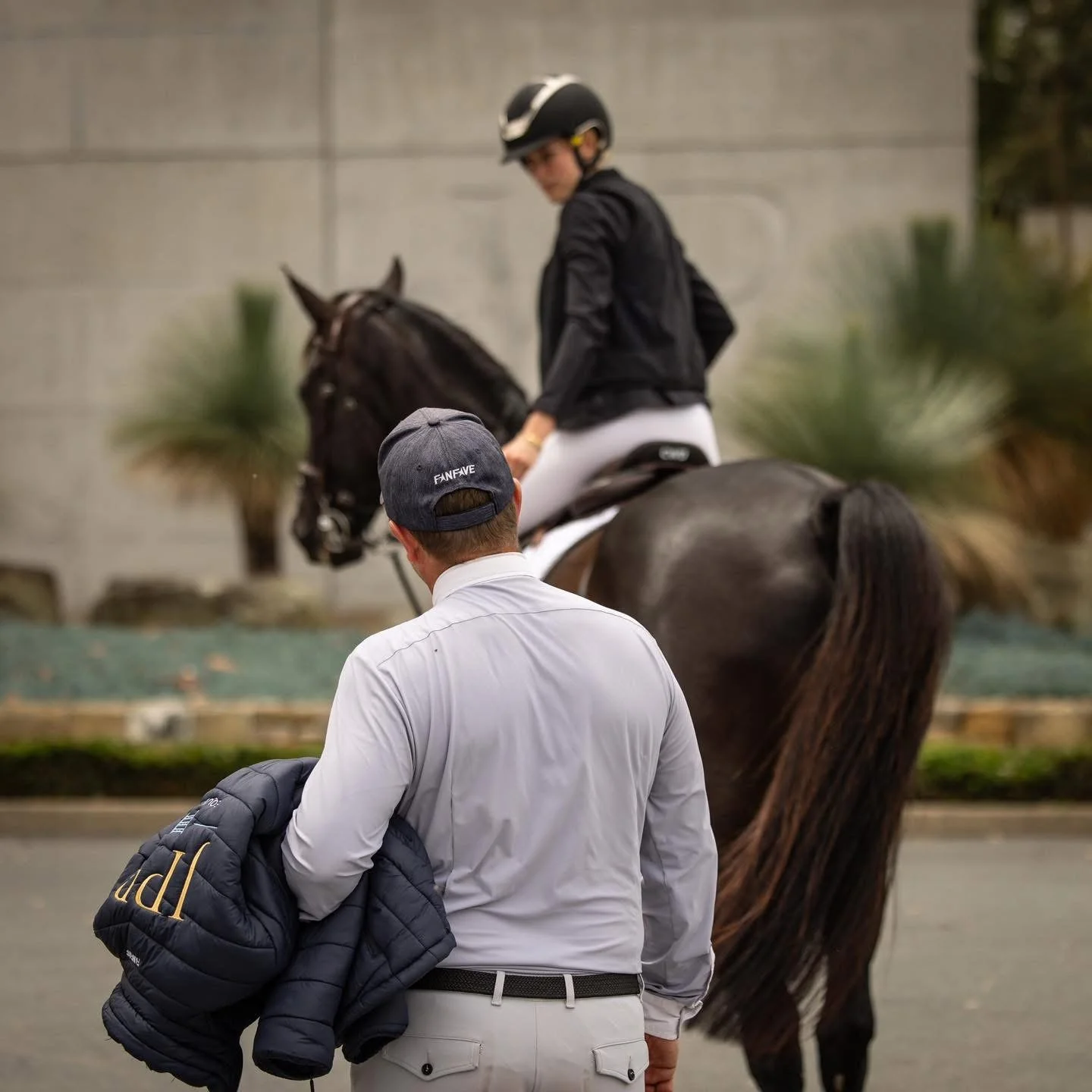 A man in a light-colored shirt and cap stands with his back to the camera, holding a jacket, as a young female equestrian rider on a dark horse rides past in an outdoor setting.
