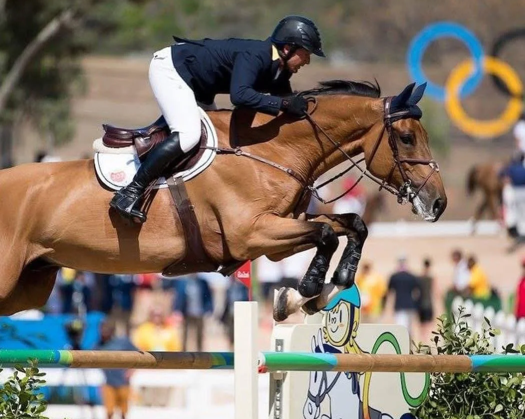 A male equestrian rider in black helmet, white breeches, and navy jacket jumping over a hurdle on a brown horse during a show jumping event, with Olympic rings visible in the background.