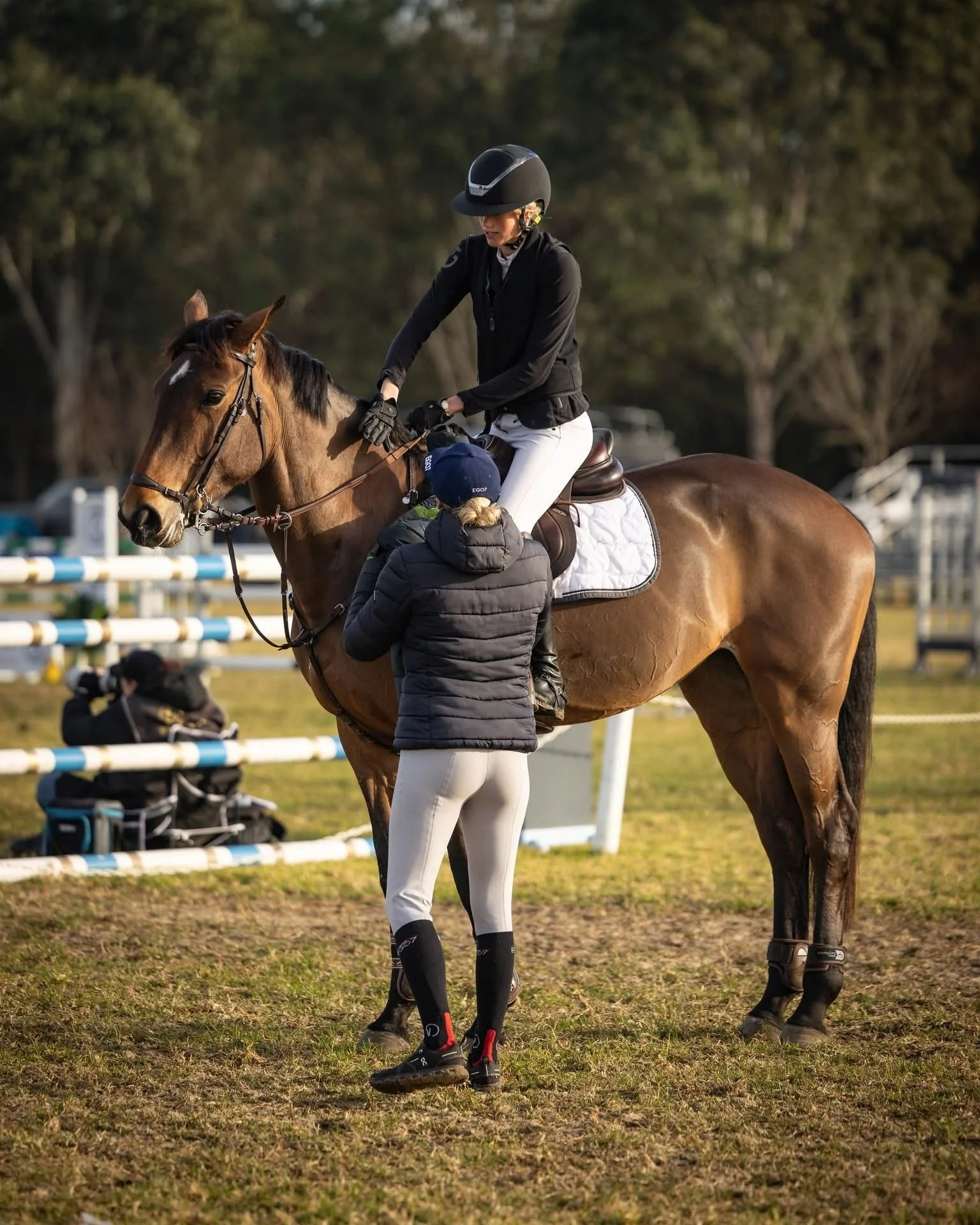 A woman in riding gear sitting on a brown horse while a instructor adjusts her saddle on an outdoor riding arena.