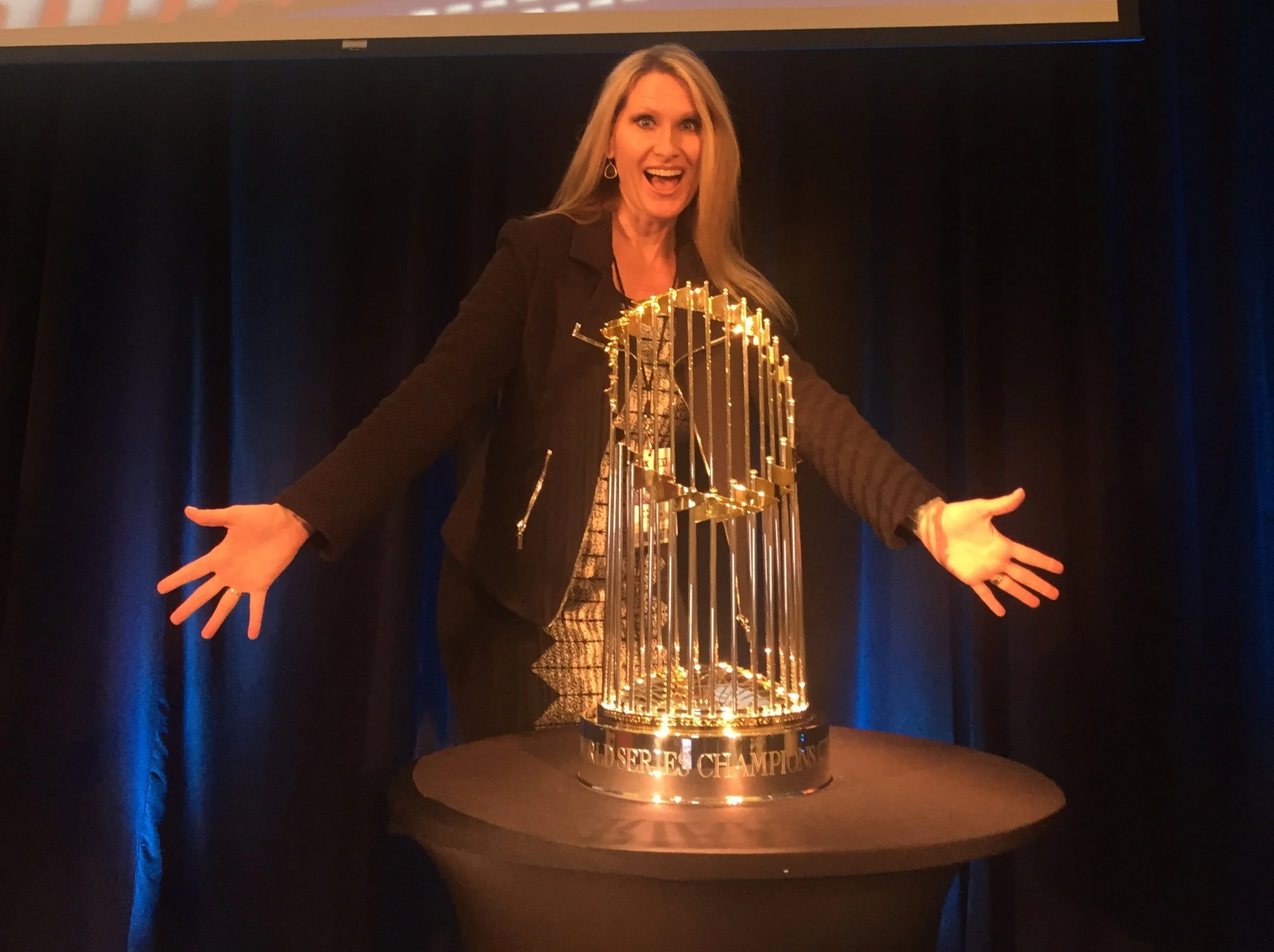 Woman celebrating with a trophy cup that has gold vertical bars and a gold circular top, standing on a black table with a black curtain background.