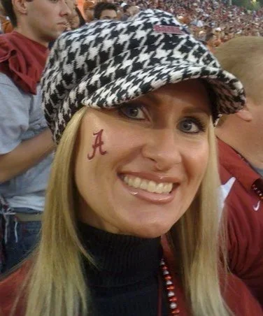 A woman wearing a black and white patterned hat with a red letter 'A' painted on her cheek, smiling at a sports event with a crowd in the background.