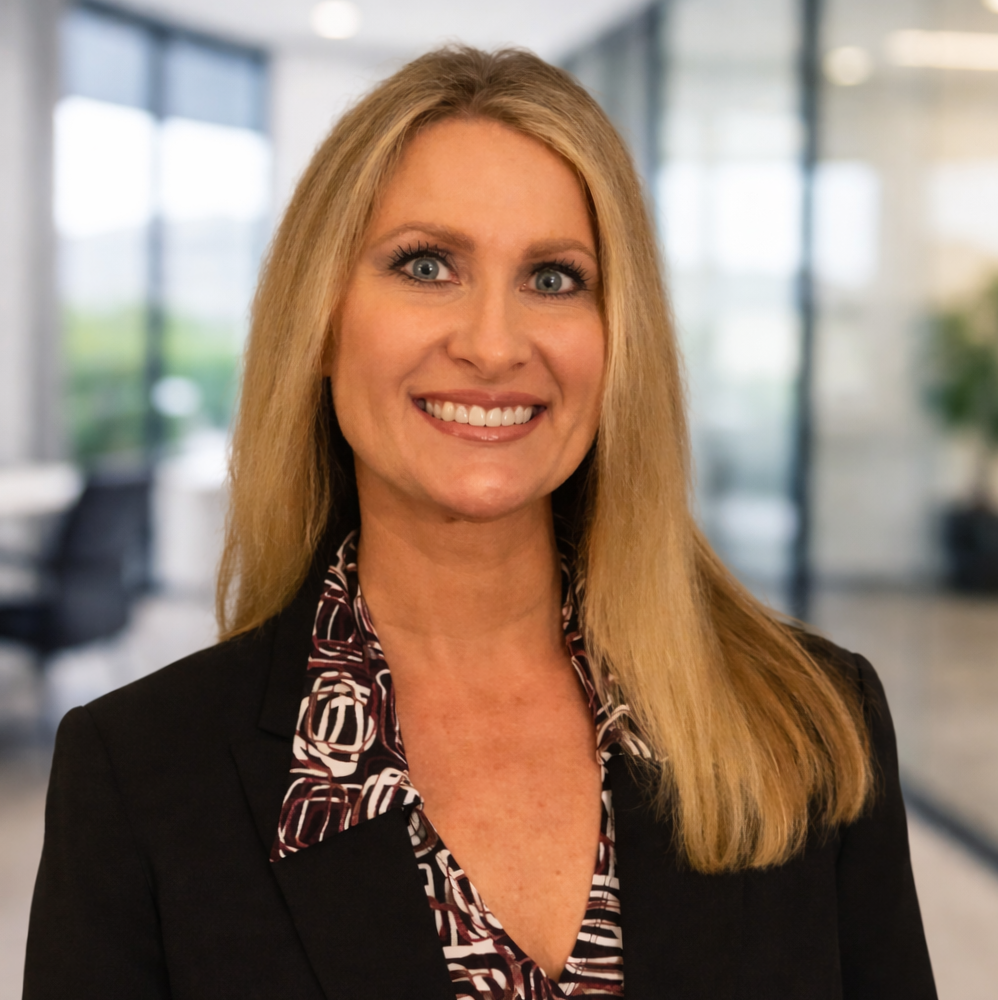 A smiling woman with long blonde hair wearing a black blazer and patterned blouse in an office setting.