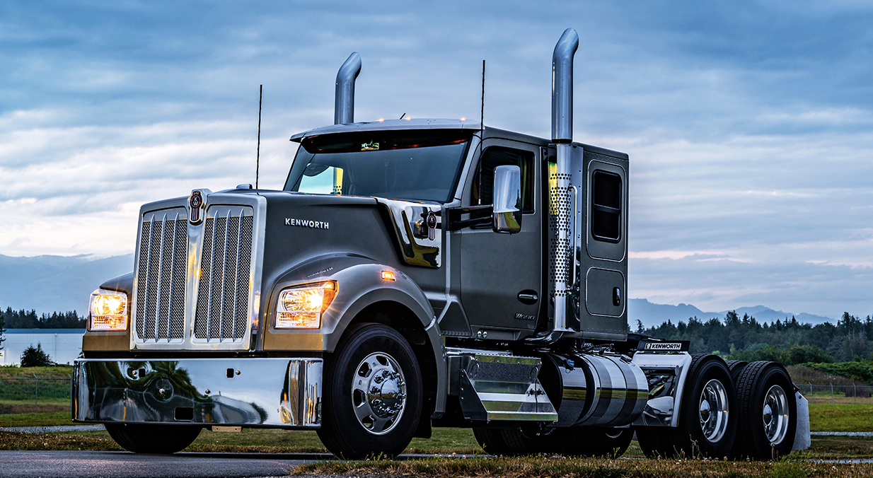 A large black semi-truck parked outdoors with mountains and a cloudy sky in the background.