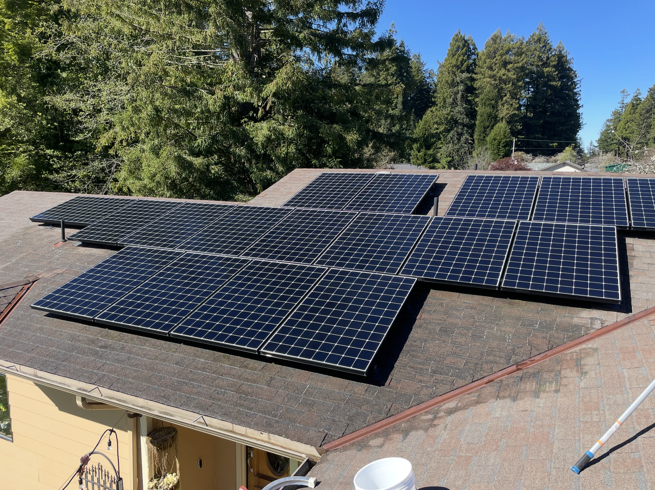 Multiple black solar panels installed on a residential roof with a background of green trees and a clear blue sky.