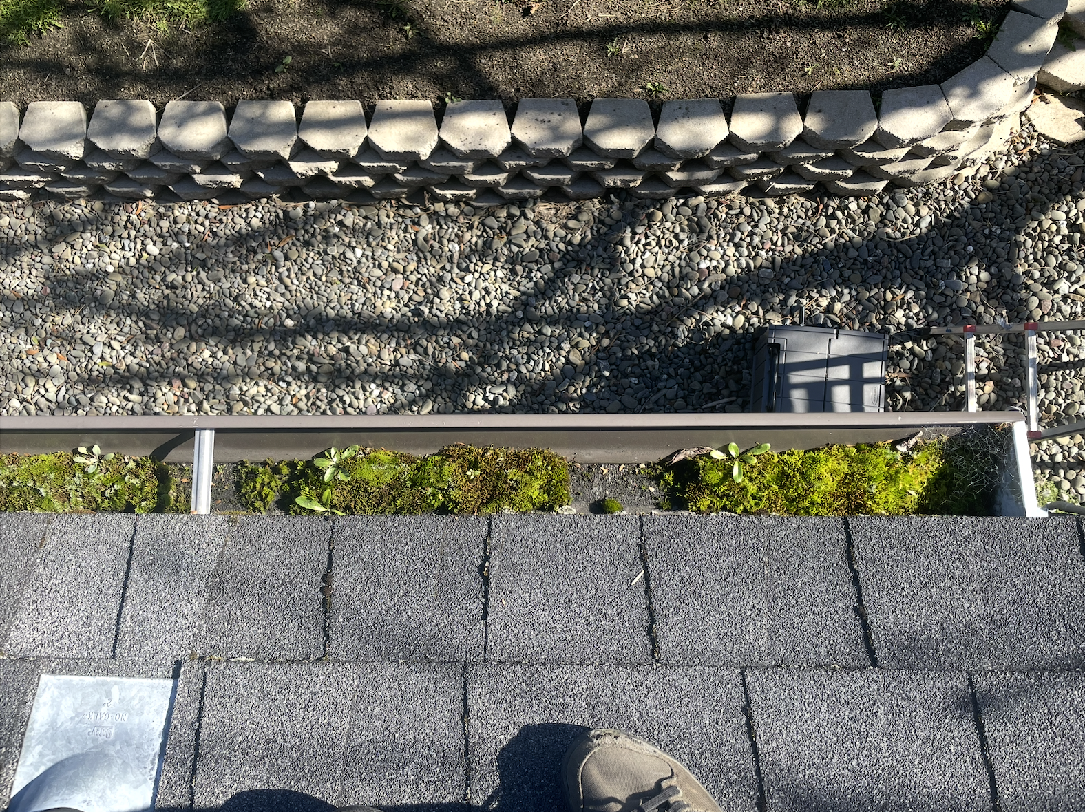 Looking down at the edge of a roof with shingles, above a small balcony or ledge with patches of moss and small plants. Below is a gravel yard with a black storage box, a ladder, and a stone border with rounded stones and soil behind it.