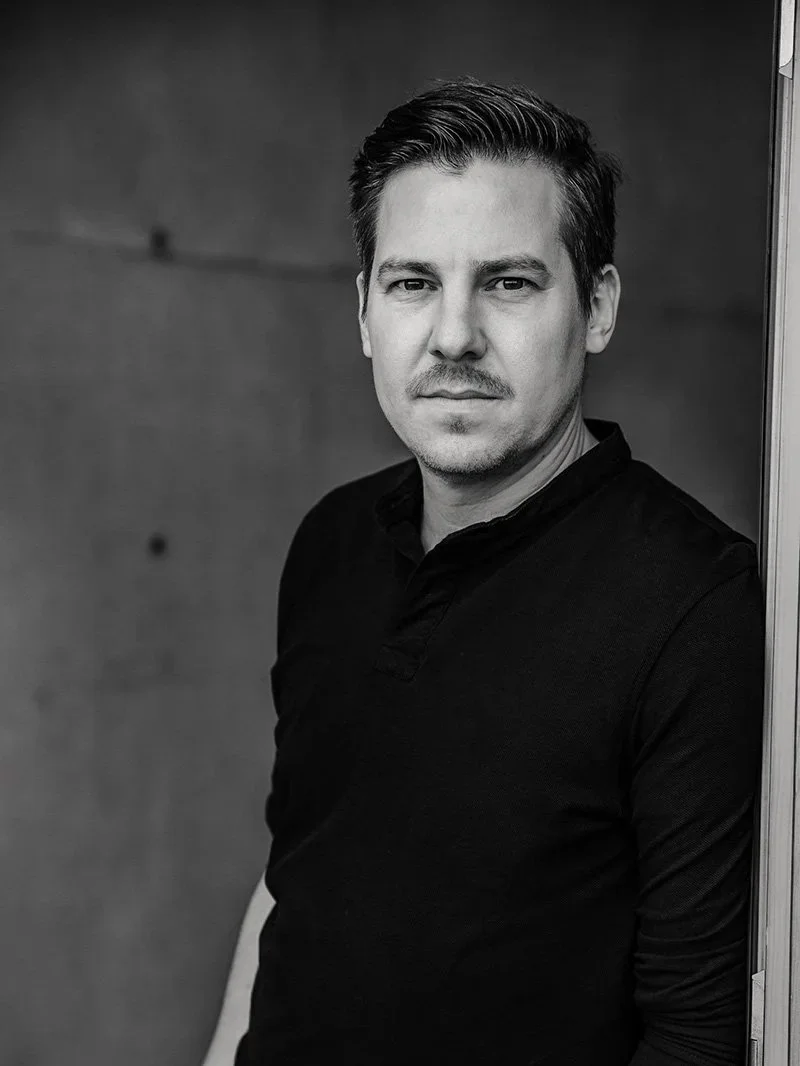 Schwarz-Weiß-Foto eines Mannes, der an einer Wand lehnt, mit schwarzem Hemd, mit ernstem Blick, dunklem Hintergrund.