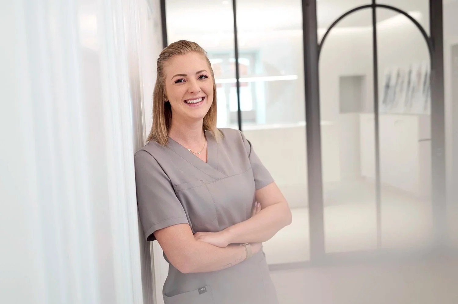 Eine lächelnde junge Frau in grauer Arbeitskleidung steht in einem modernen, gut beleuchteten Bürogebäude, lehnt sich an eine weiße Wand und hat die Arme verschränkt.