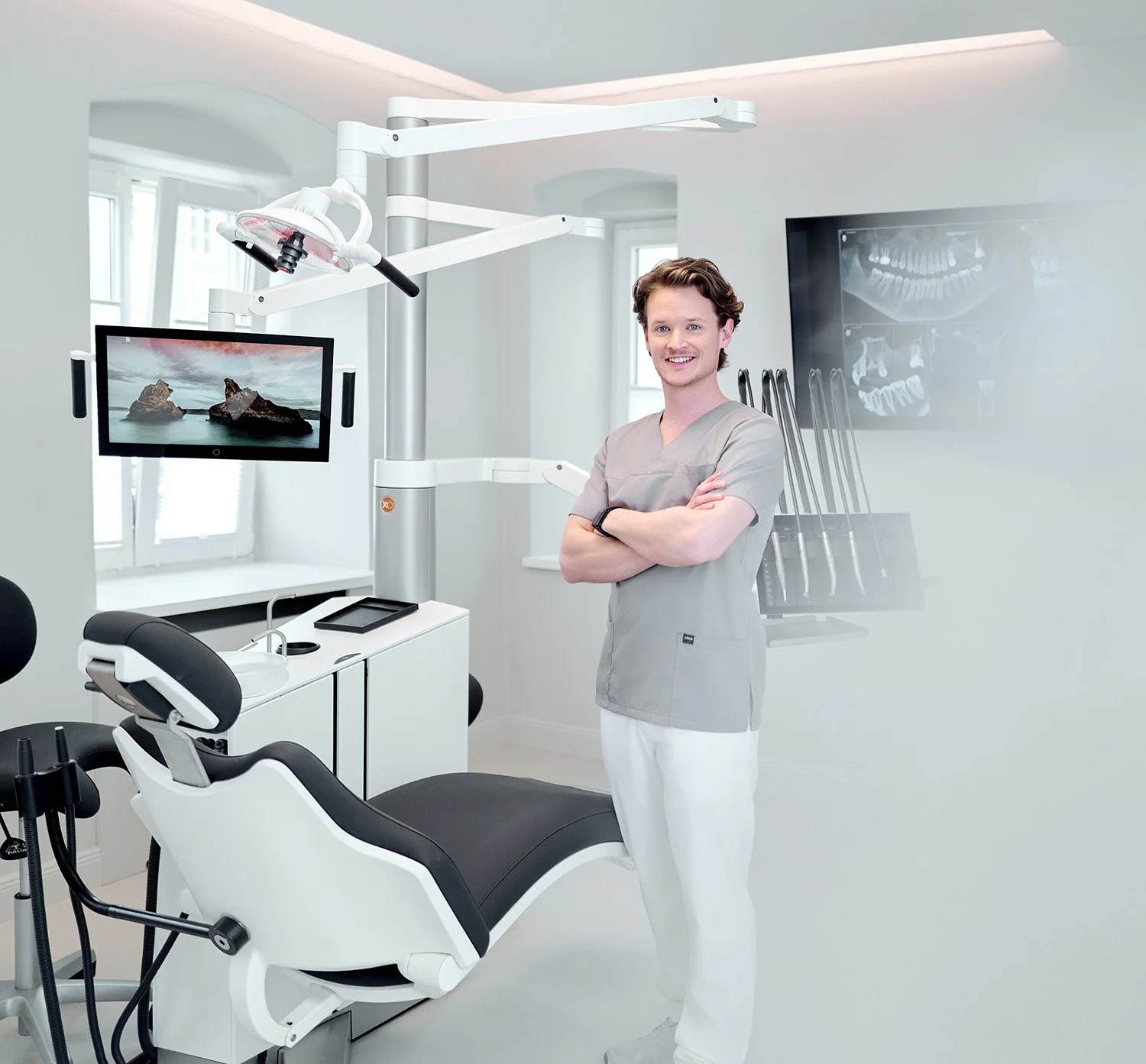 Ein zahnärztlicher Behandlungsraum mit einem lachenden männlichen Zahnarzt in einem grauen Arztkittel, der vor einem zahnärztlichen Behandlungsstuhl steht. Es sind moderne Dentalgeräte, ein Monitor und eine Wand mit Röntgenaufnahmen sichtbar.