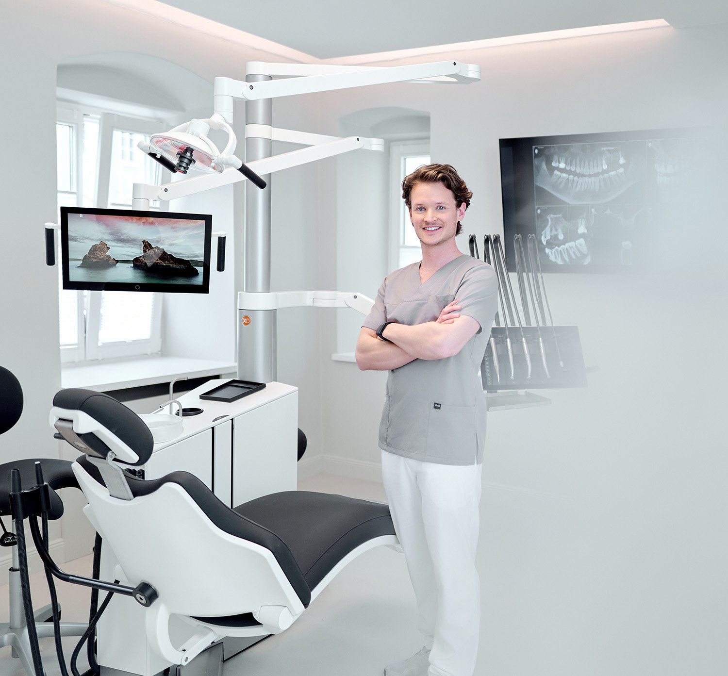 Ein zahnärztlicher Behandlungsraum mit einem lachenden männlichen Zahnarzt in einem grauen Arztkittel, der vor einem zahnärztlichen Behandlungsstuhl steht. Es sind moderne Dentalgeräte, ein Monitor und eine Wand mit Röntgenaufnahmen sichtbar.