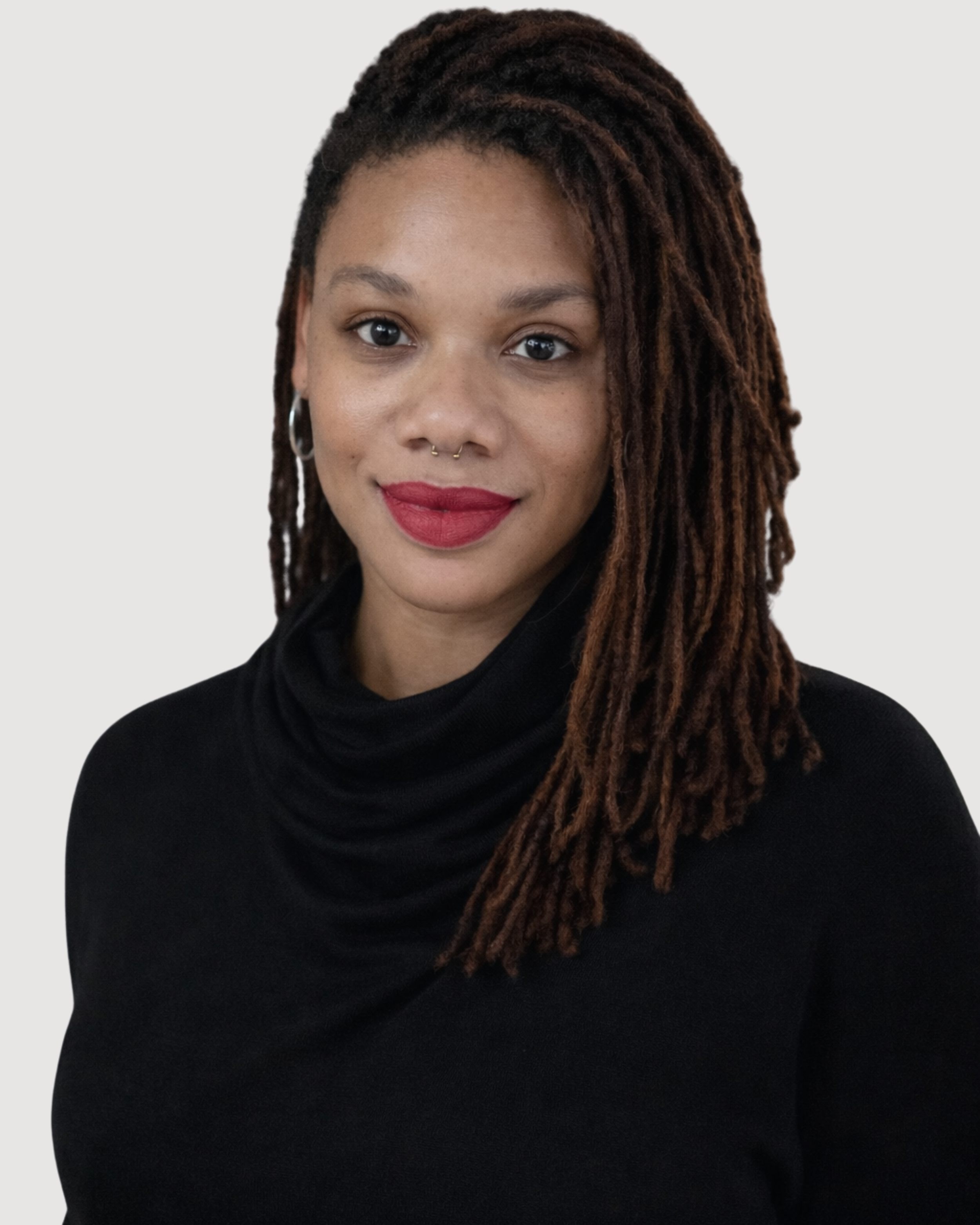 Portrait of a woman with brown dreadlocks, wearing a black turtleneck and red lipstick, smiling gently against a plain light background.
