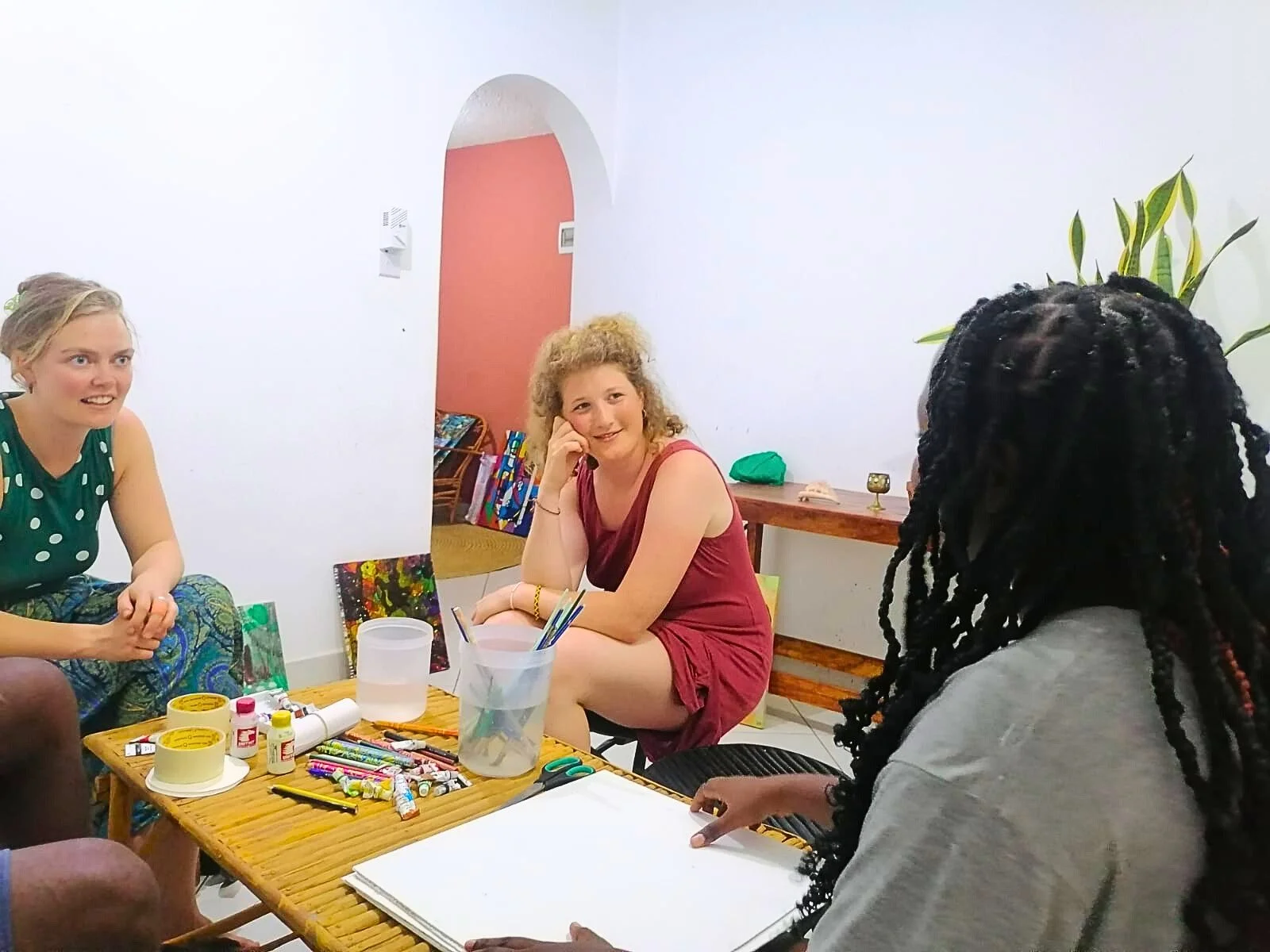 Three women sitting around a table with art supplies, engaging in a conversation in a casual indoor setting.