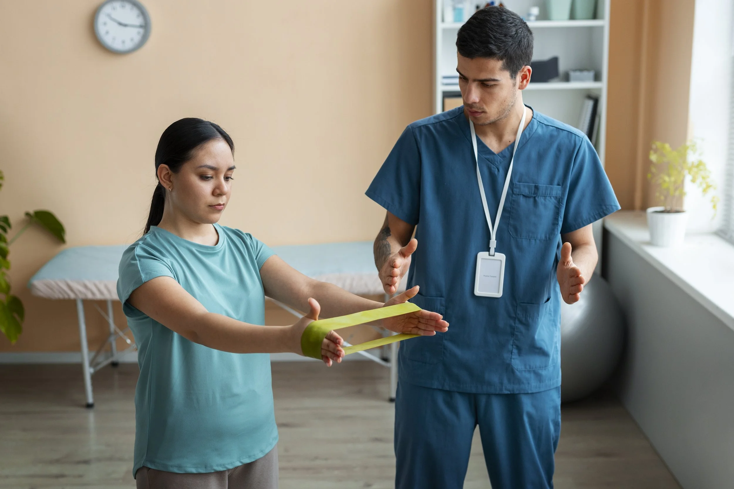 A female patient in a teal shirt with her arm extended and a healthcare worker in blue scrubs explaining something in a medical setting.