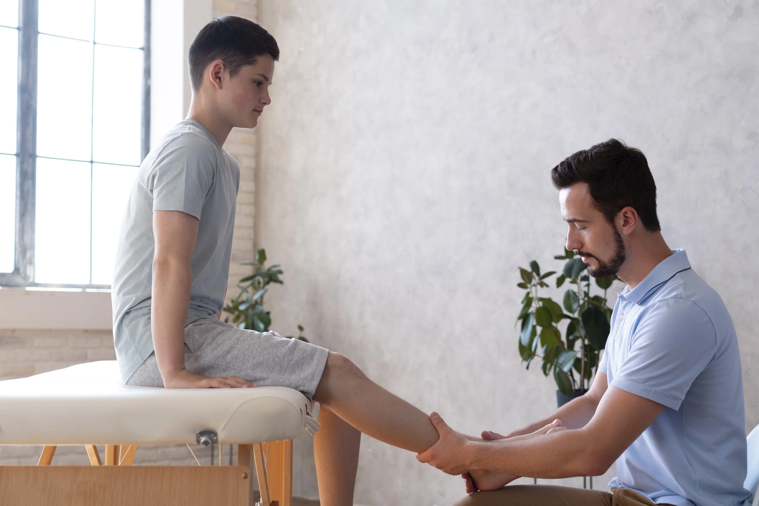 A young woman sitting on an examination table with her leg extended, being examined by a male healthcare professional in a clinical setting with large windows and plants in the background.