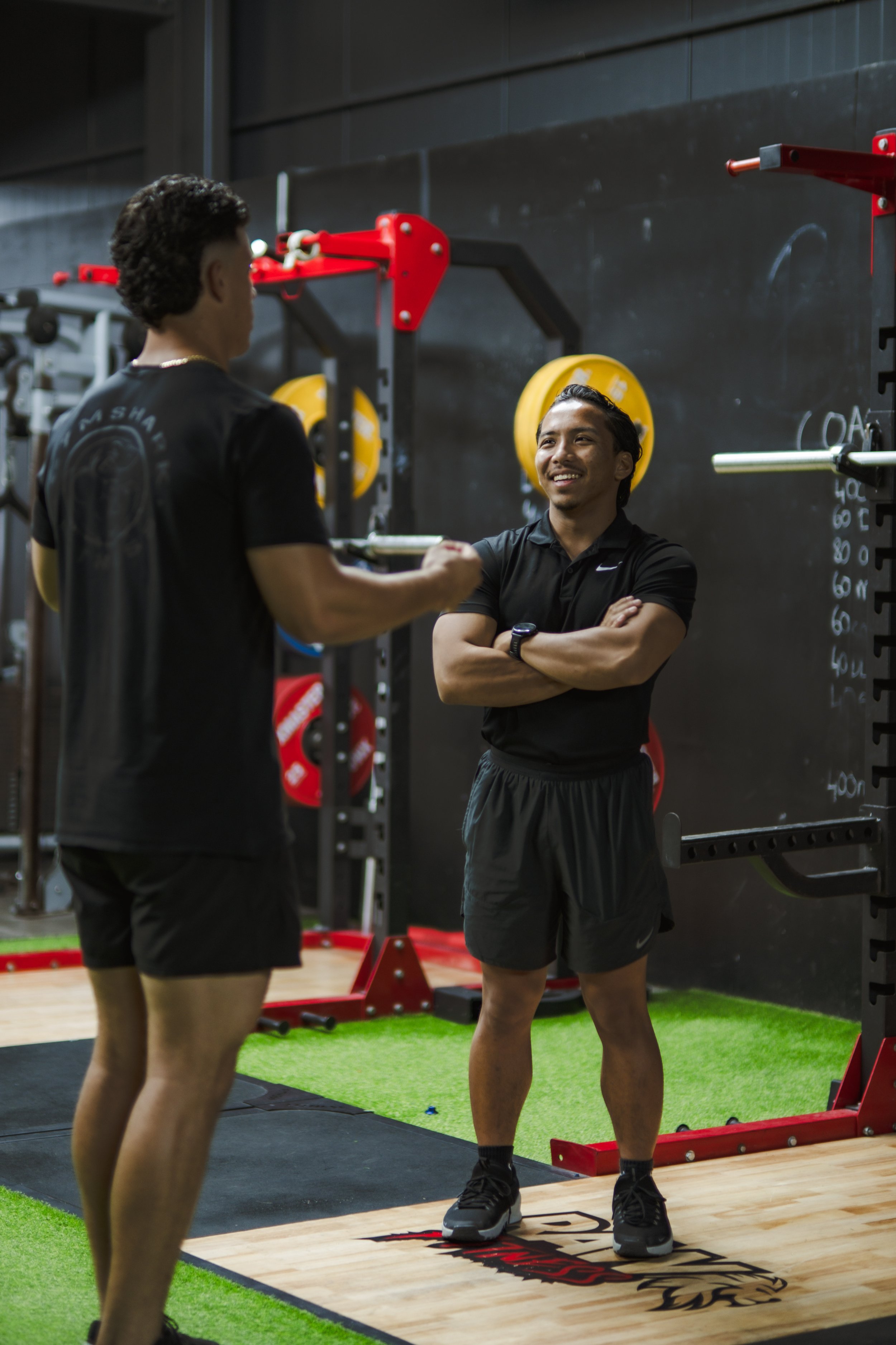 Two men in a gym having a conversation. The man on the right is smiling with arms crossed, wearing black athletic clothes and sneakers. The man on the left, seen from behind, is also in black athletic clothes. The background features gym equipment, weights, and a blackboard.