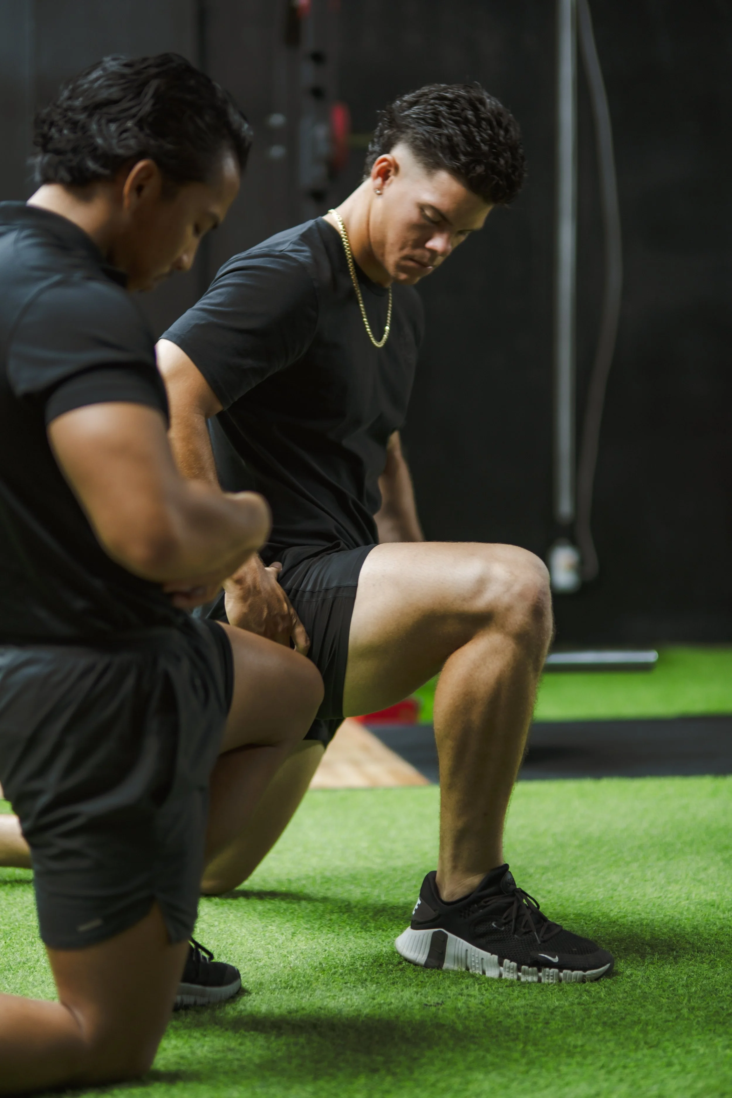 Two young men are kneeling on green gym flooring, adjusting their athletic shoes before exercising, with a dark gym background.