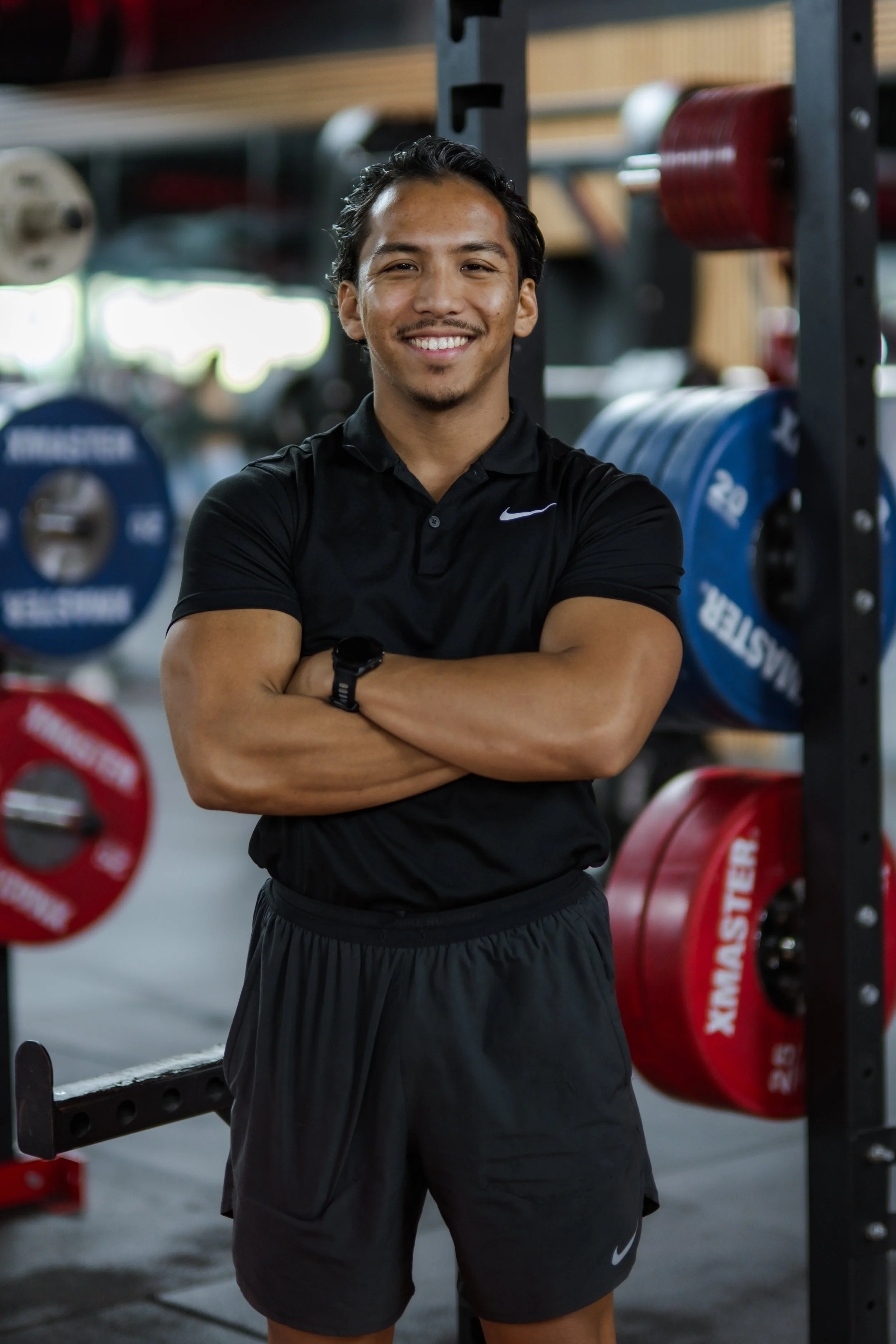 Young man smiling, with arms crossed, standing in a gym with weightlifting equipment and colored weight plates in the background.
