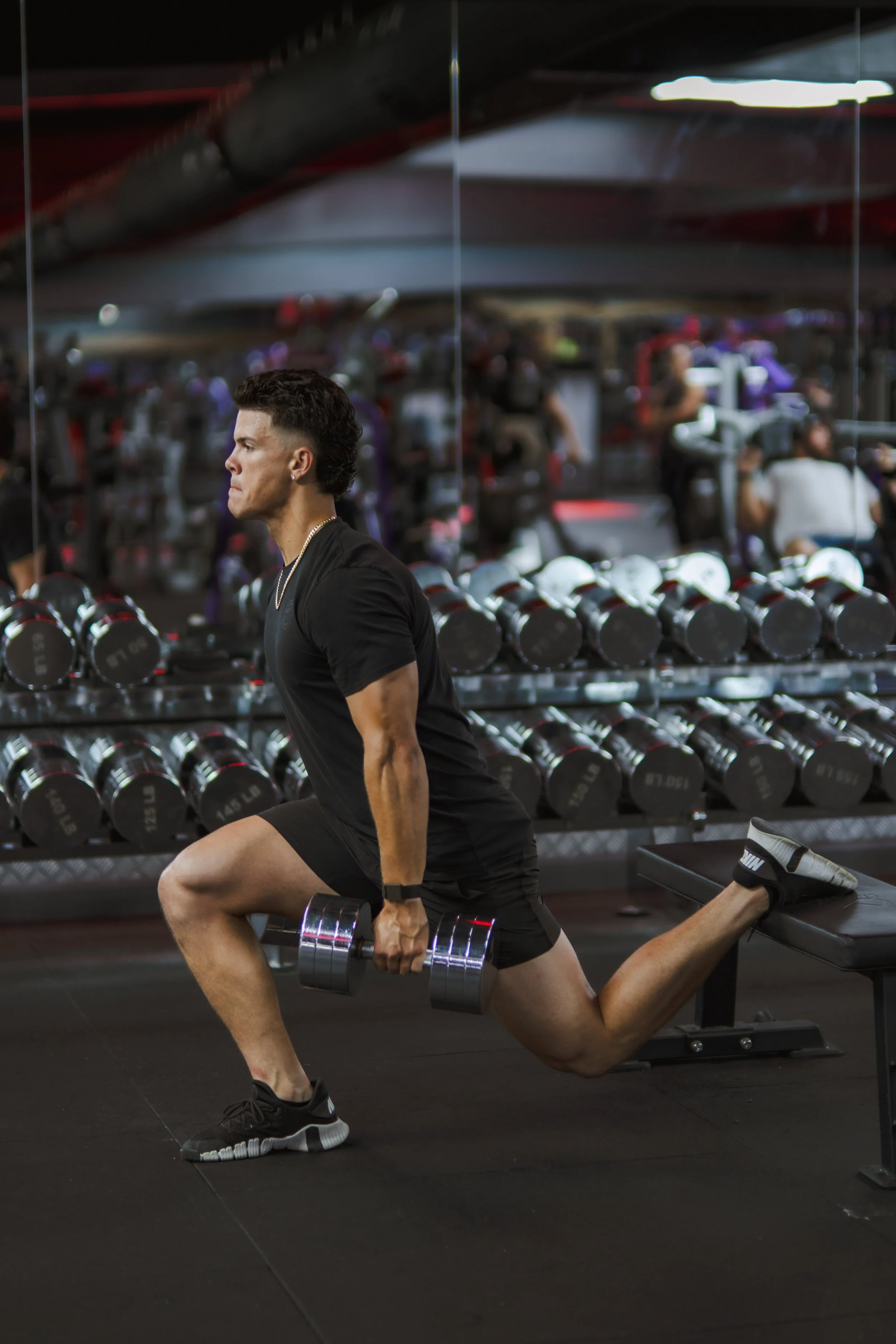 A man performing a lunge exercise with a dumbbell in his right hand in a gym, with weight racks and gym equipment blurred in the background.