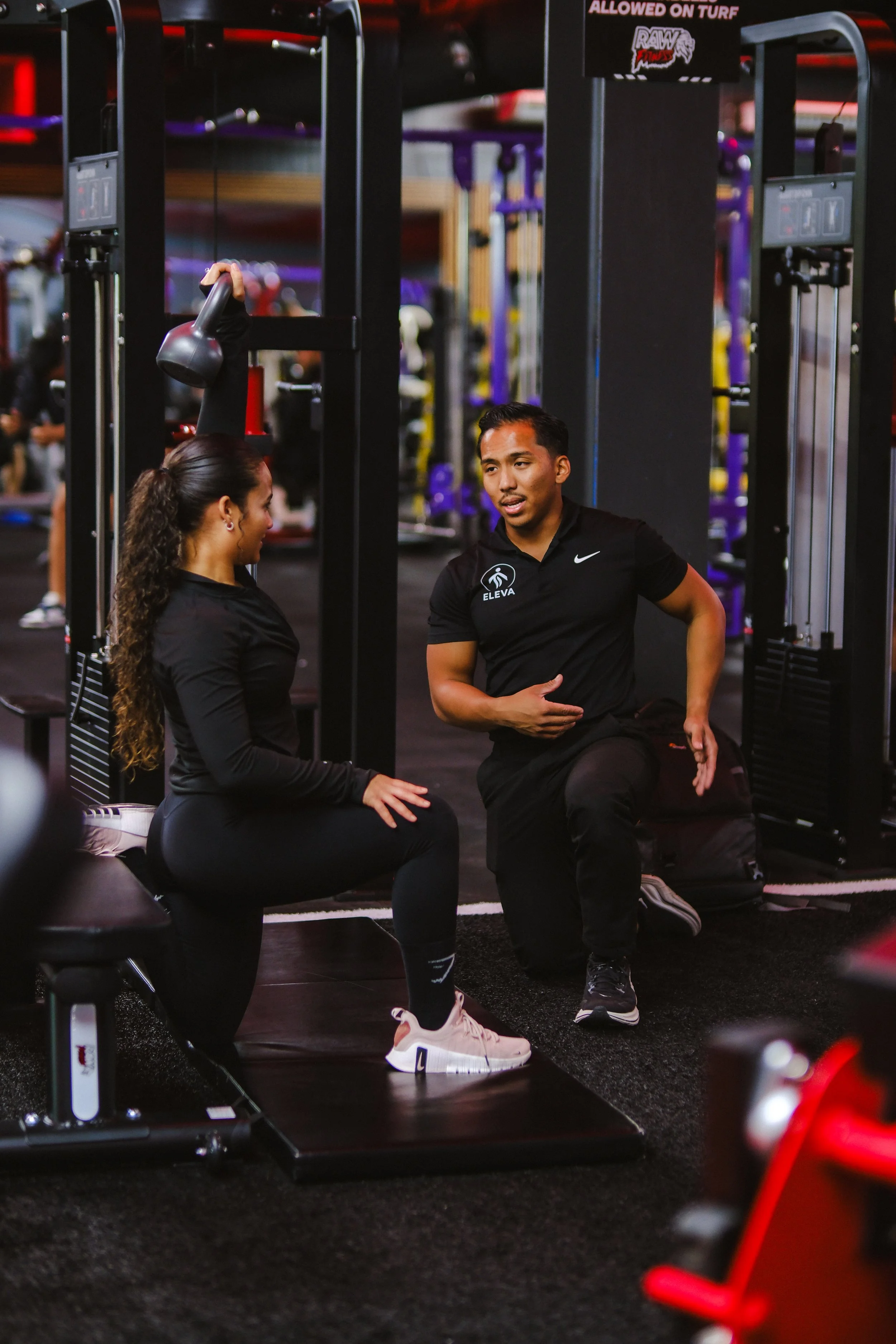A man and a woman having a conversation in a gym, with workout equipment and a black floor visible.