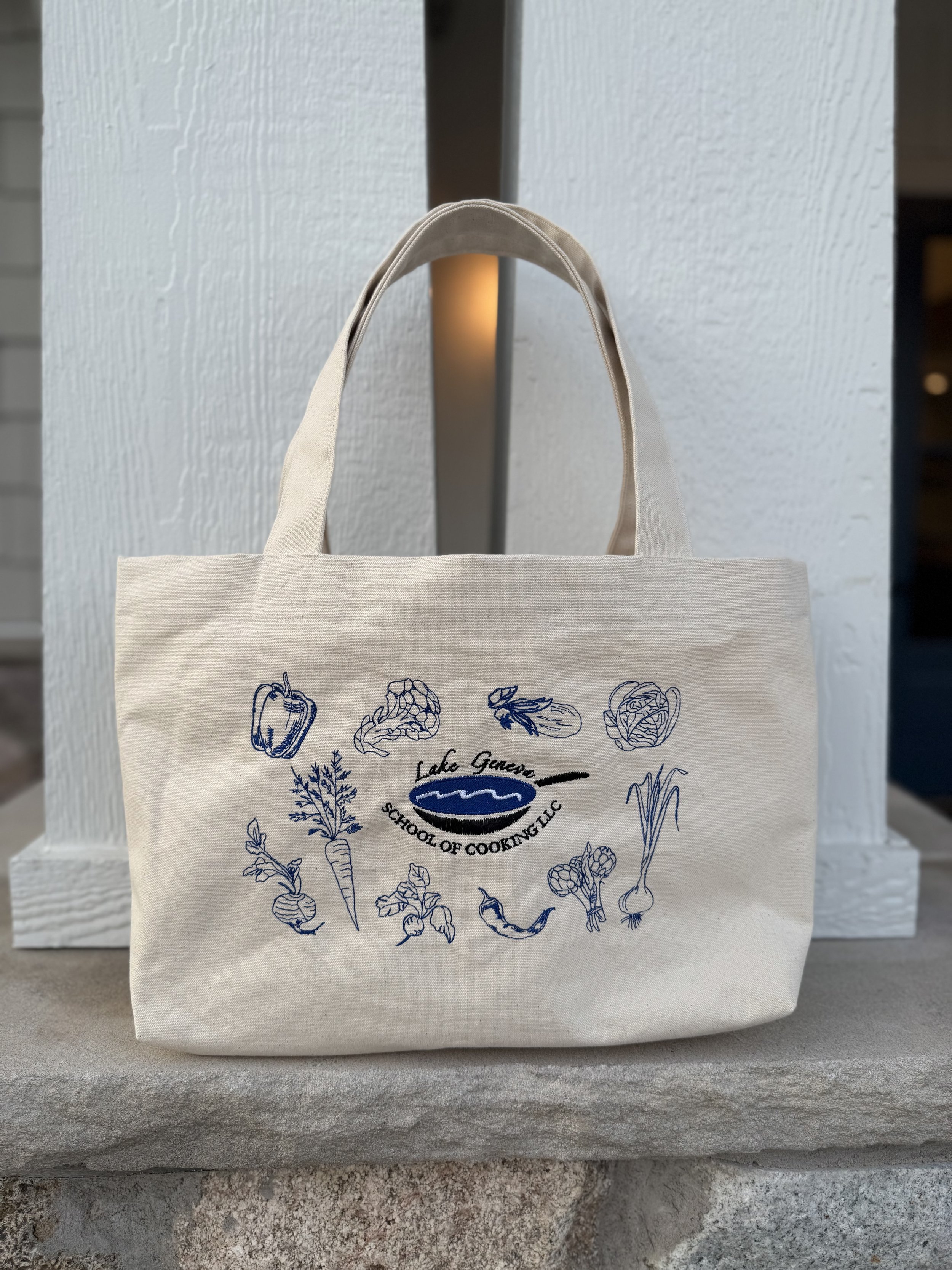 A beige tote bag with blue sketches of vegetables and herbs, featuring a logo for Lake Geneva School of Cooking LLC.