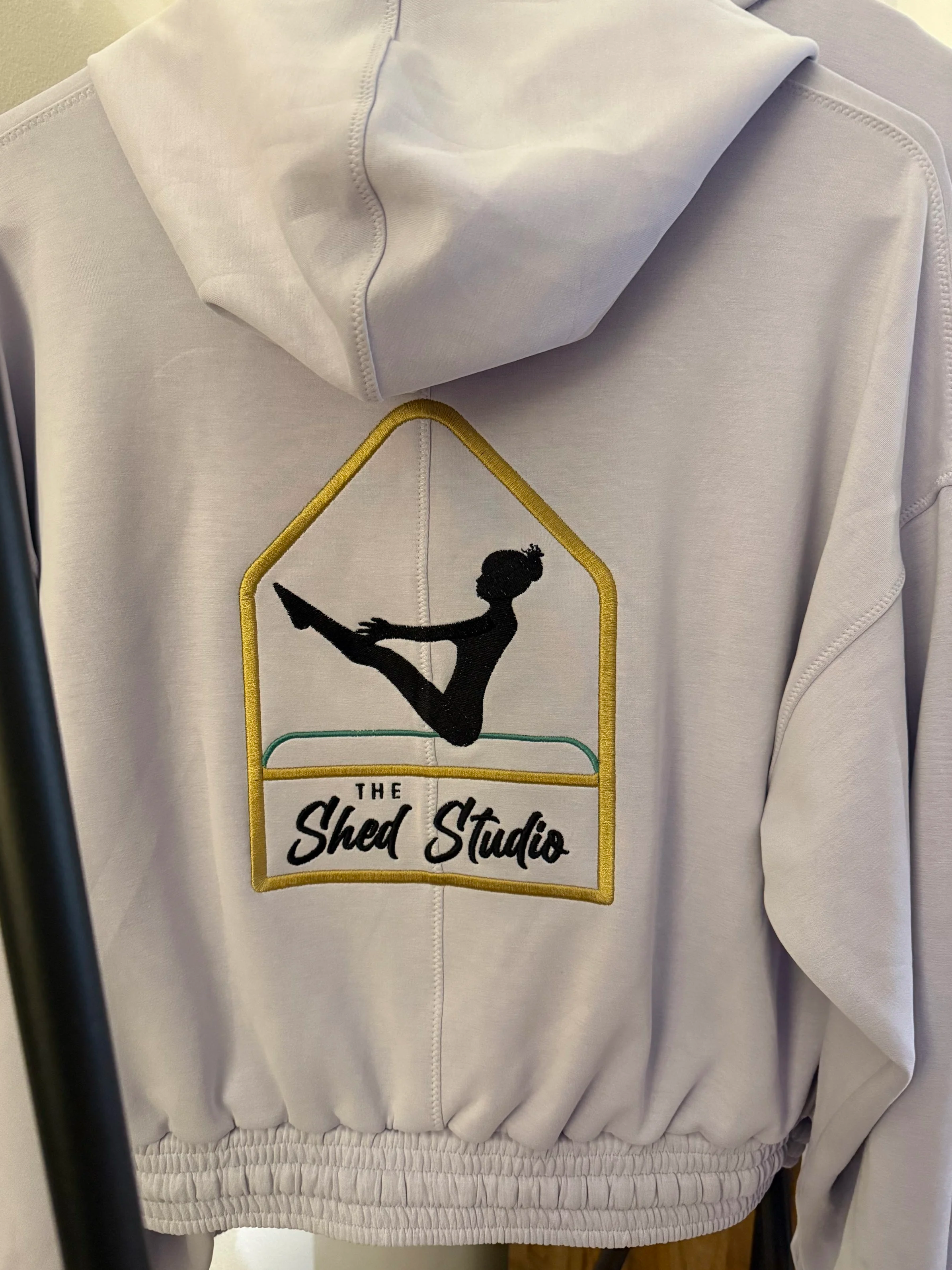 White hoodie with a patch featuring a silhouette of a woman doing a split, with the text "The Shed Studio" underneath, on the back of the hoodie.