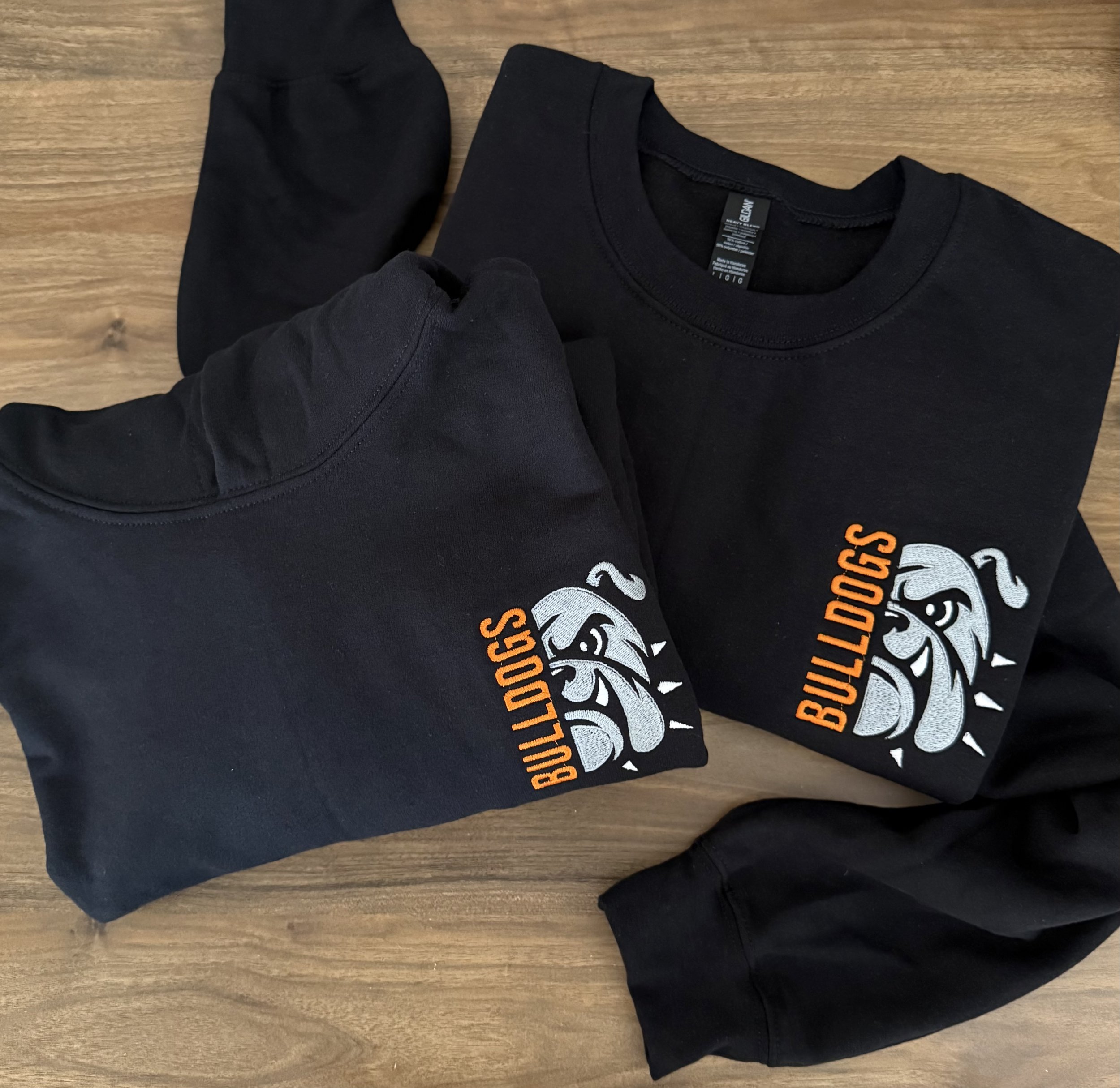 Two black T-shirts with Bulldog logo and the word "Bulldogs" in orange, laid out on a wooden surface, one with sleeves folded.