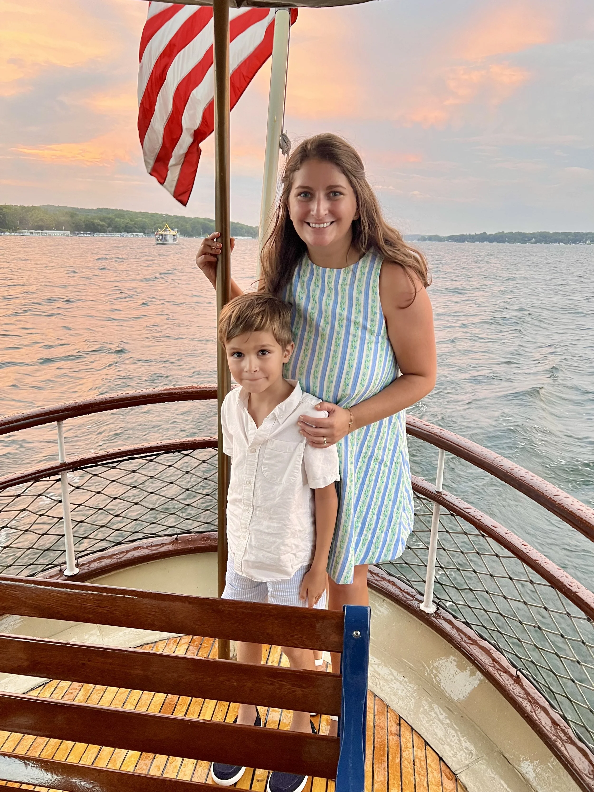 A woman and a young boy standing on a boat with an American flag, with a body of water and a sunset sky in the background.