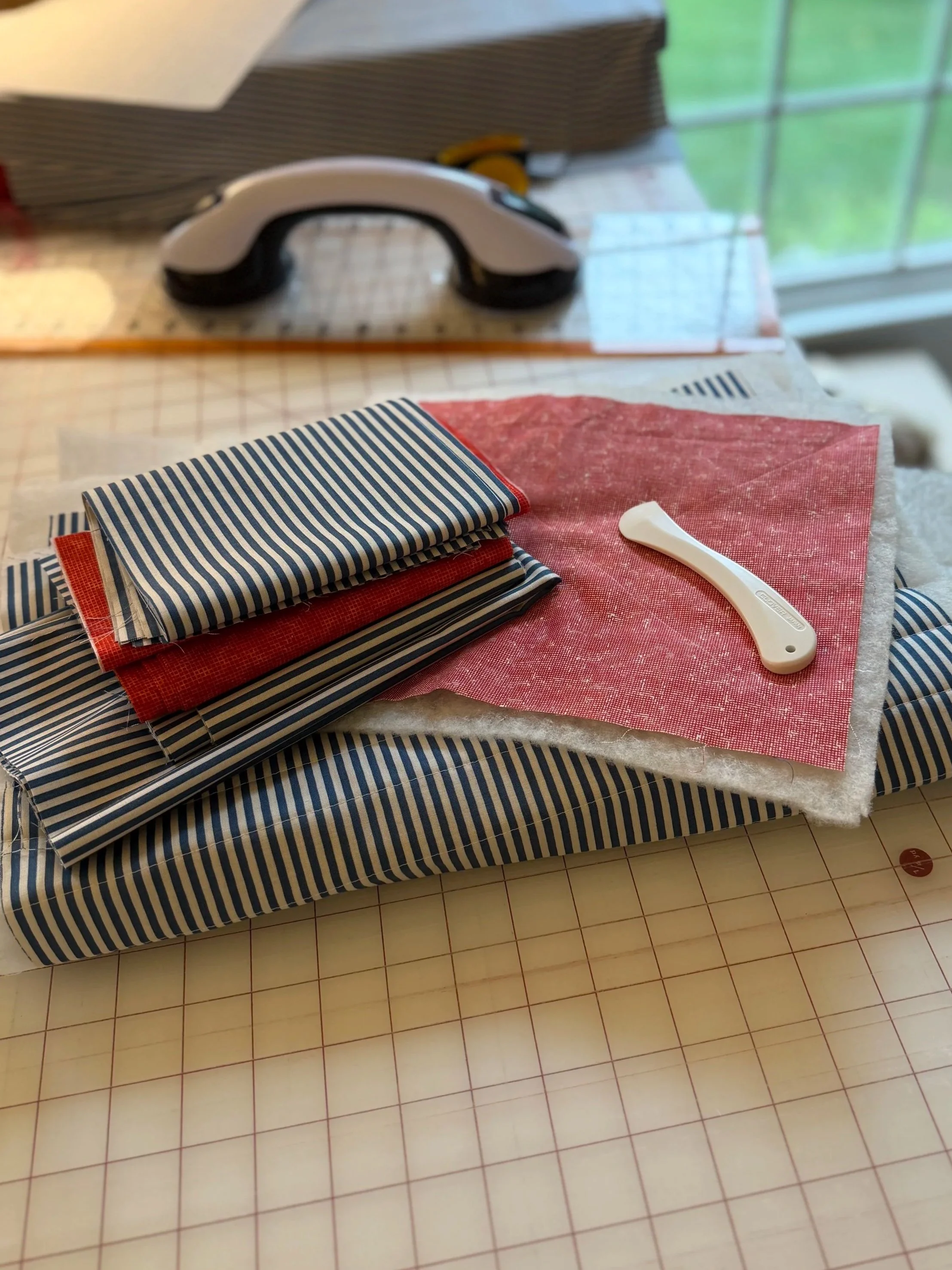 Sewing supplies including folded striped fabric, red fabric, a white seam ripper, and a pink textured fabric on a table with a cutting mat, sewing goggles above, and a window in the background.
