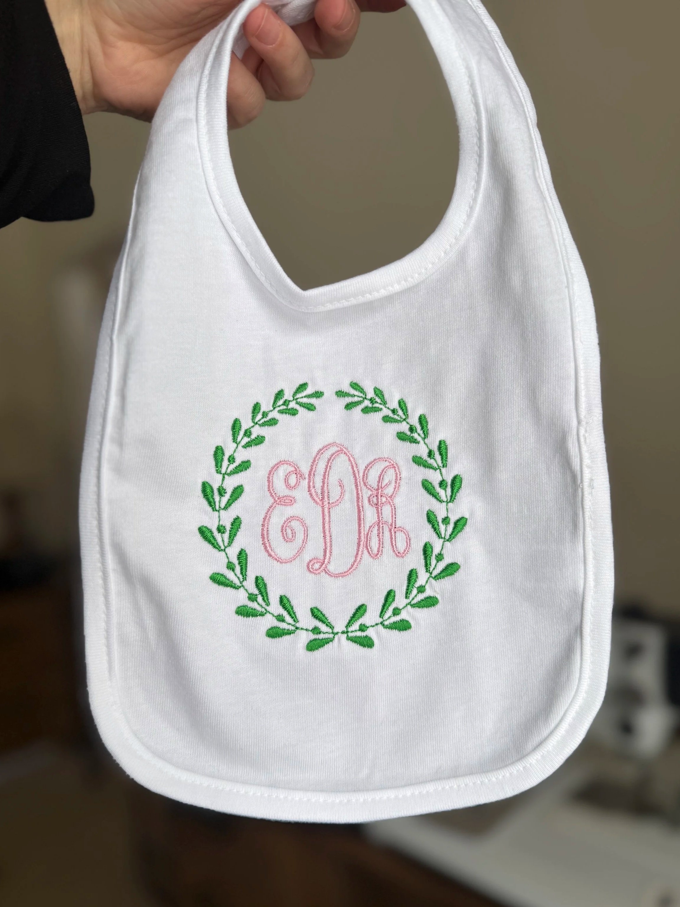 White baby bib with green embroidered laurel wreath and pink embroidered letter 'E' at the center.