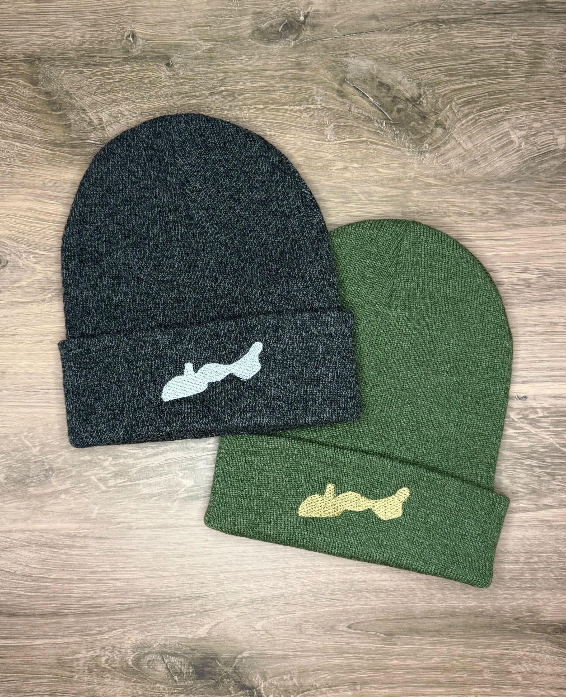 Two knit beanies, one dark gray with a white whale embroidered on the cuff and one green with a yellow whale embroidered on the cuff, placed on a wooden surface.