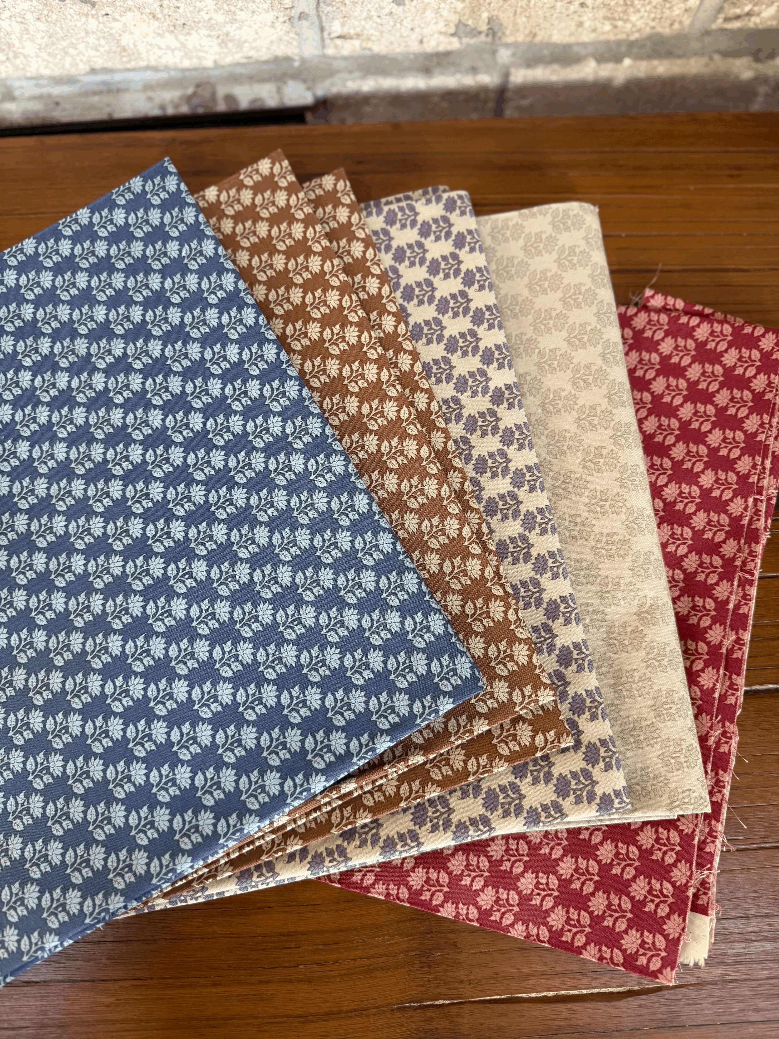 Five stacks of patterned fabric or paper in blue, brown, purple, cream, and red colors, with floral designs, laid out on a wooden surface.