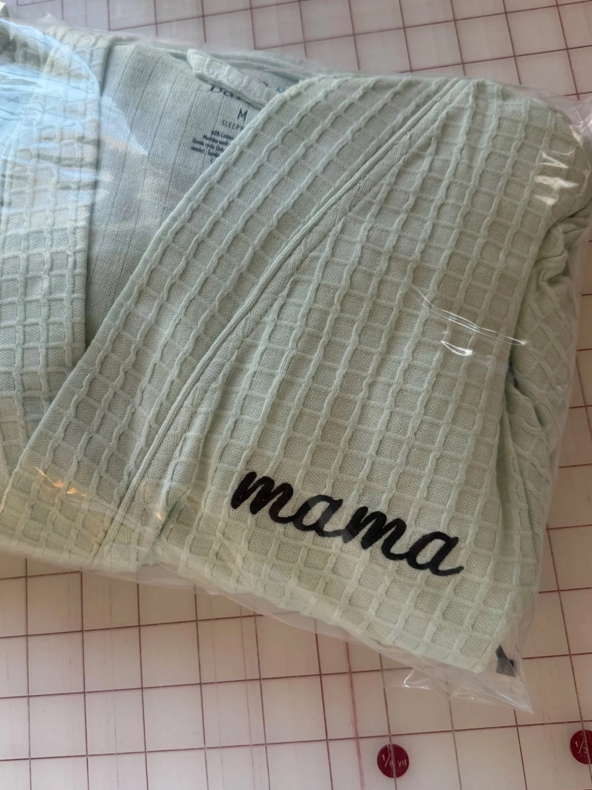 A white waffle-knit bathrobe with the word "mama" embroidered in black on the lower edge, packaged in clear plastic.