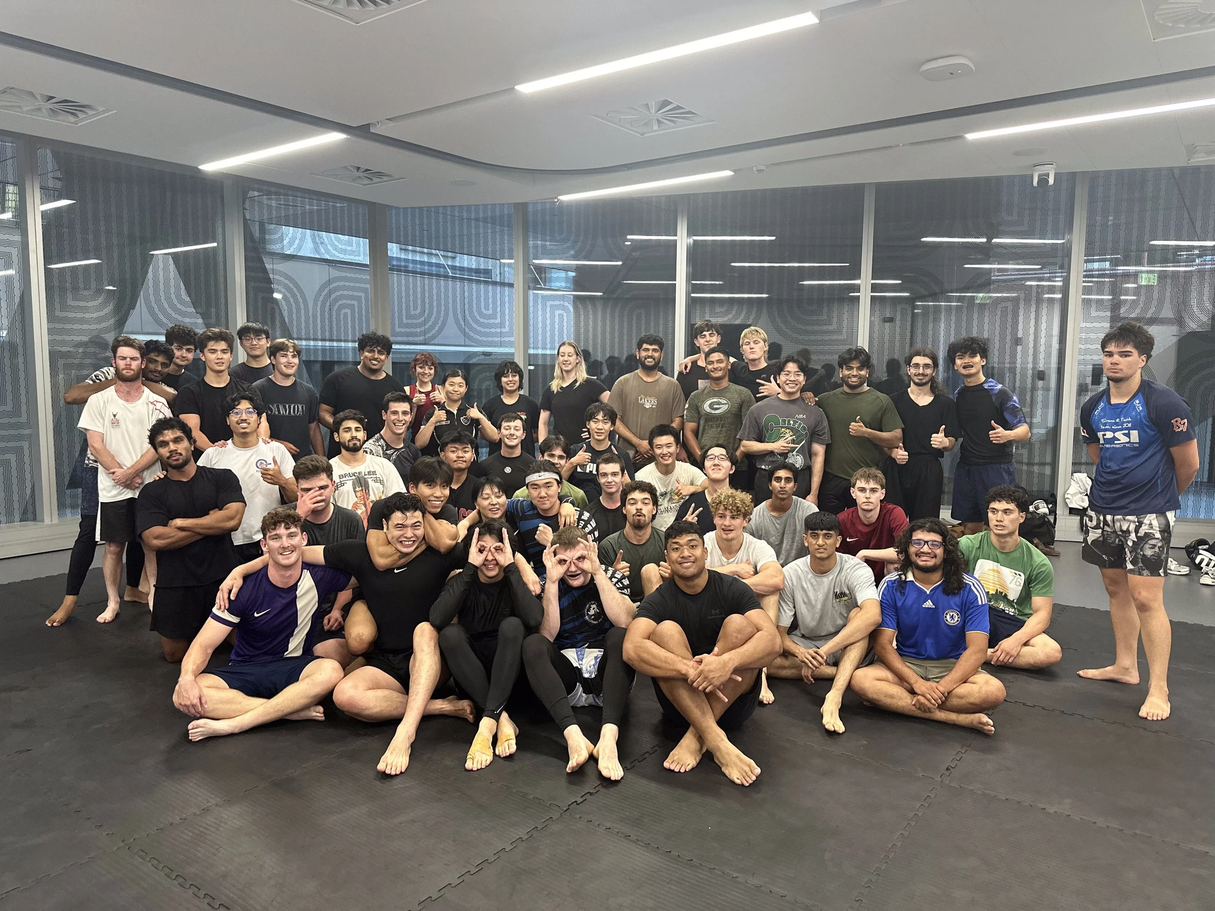 A large group of young people gathered in a sports facility, some sitting and some standing, with many smiling and making gestures. They are dressed in casual and sportswear, including some with martial arts or wrestling uniforms. The background shows a modern interior with large windows and ceiling lights.