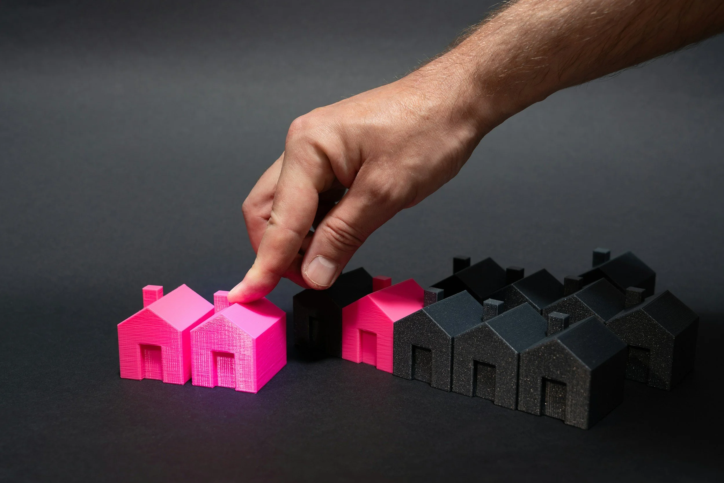 A hand placing a pink house-shaped model among other pink and black house models on a black surface.