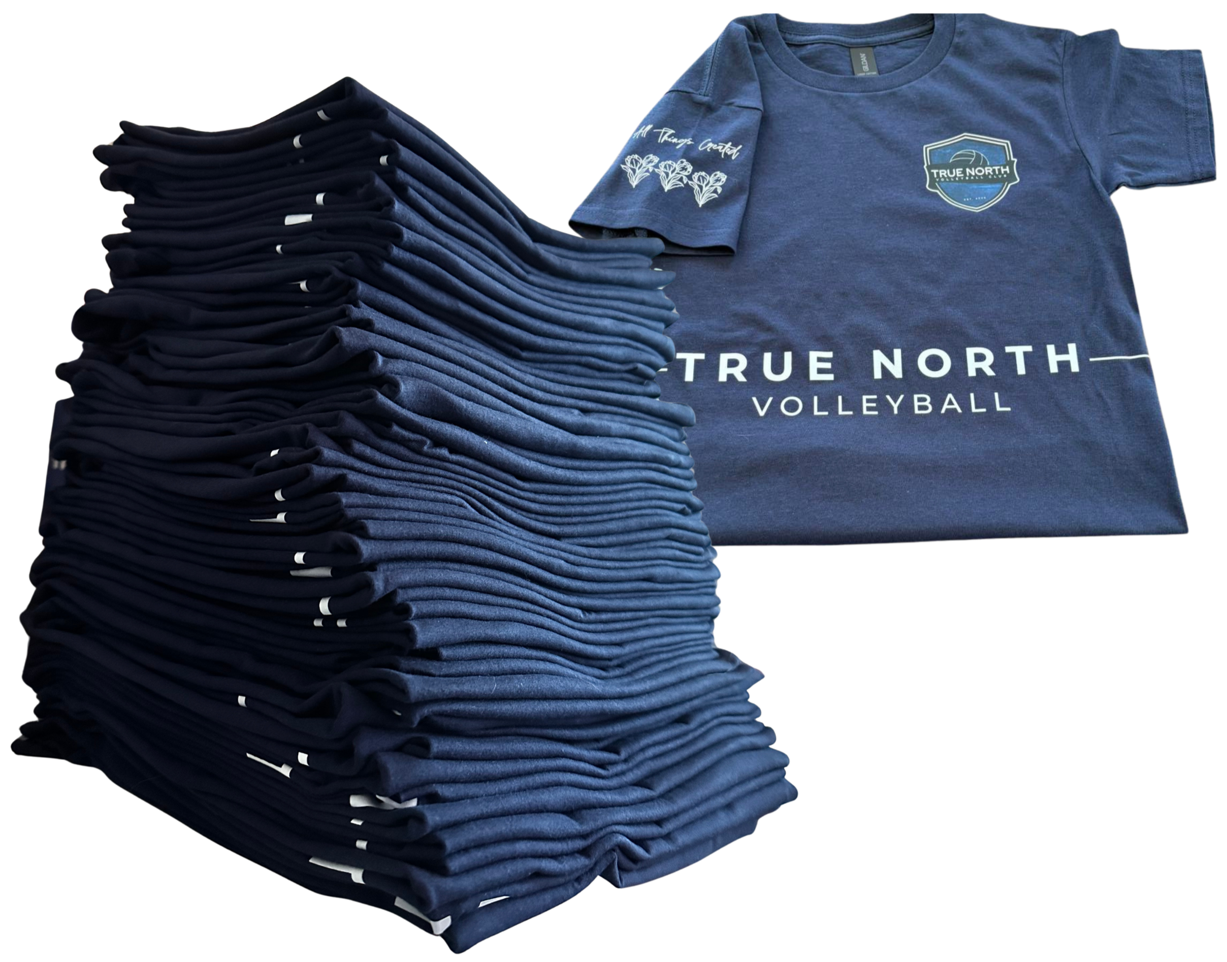 A stack of navy blue t-shirts folded neatly, with a single navy blue t-shirt laid out flat next to the stack. The laid out t-shirt has the words "TRUE NORTH VOLLEYBALL" printed on it, along with a small emblem featuring a stylized volleyball and the phrase "All Things Created" with floral illustrations.