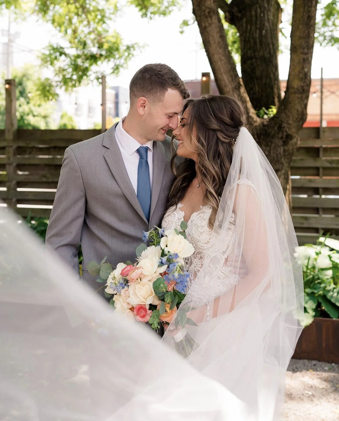 A bride and groom standing close together outdoors under a tree, holding a bouquet of flowers, with foreheads touching and smiling.