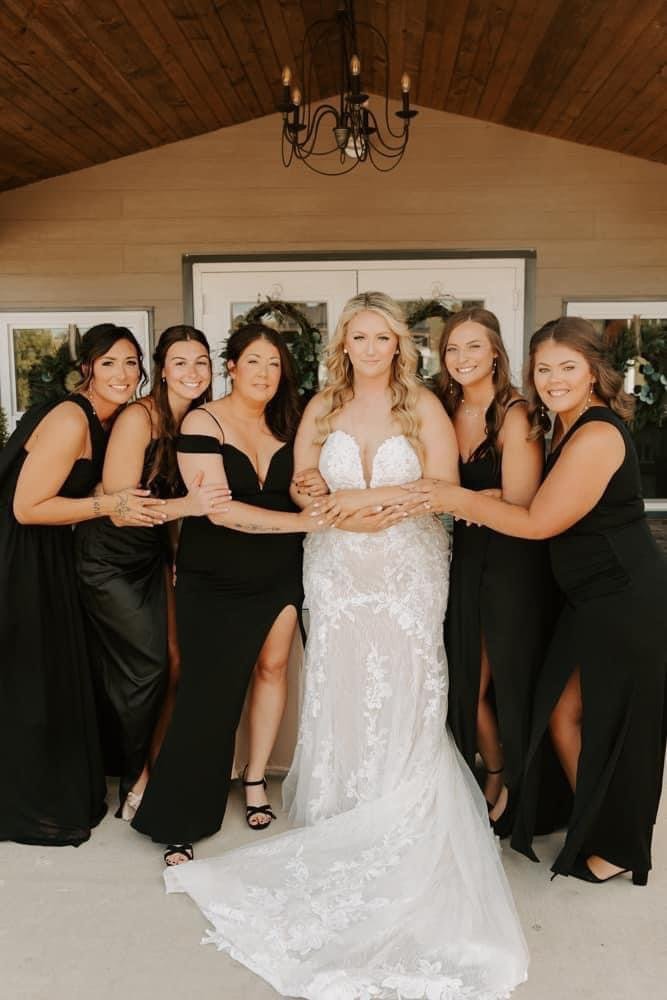 A bride in a white lace wedding gown surrounded by six bridesmaids in black dresses, standing inside a building with wood-paneled ceiling and a chandelier.