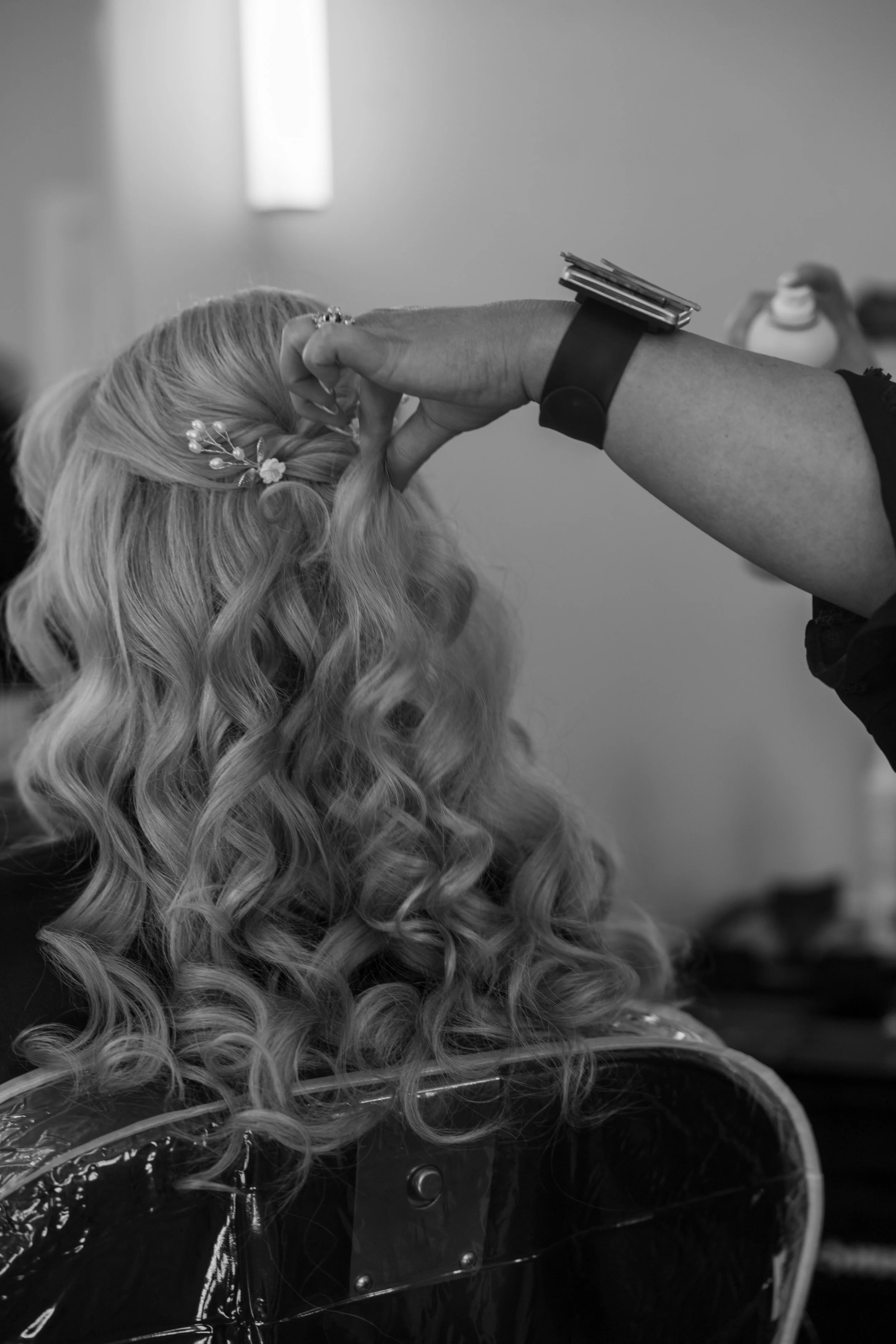 A hairstylist styles a woman's long, wavy hair with an accessory, in a salon setting.