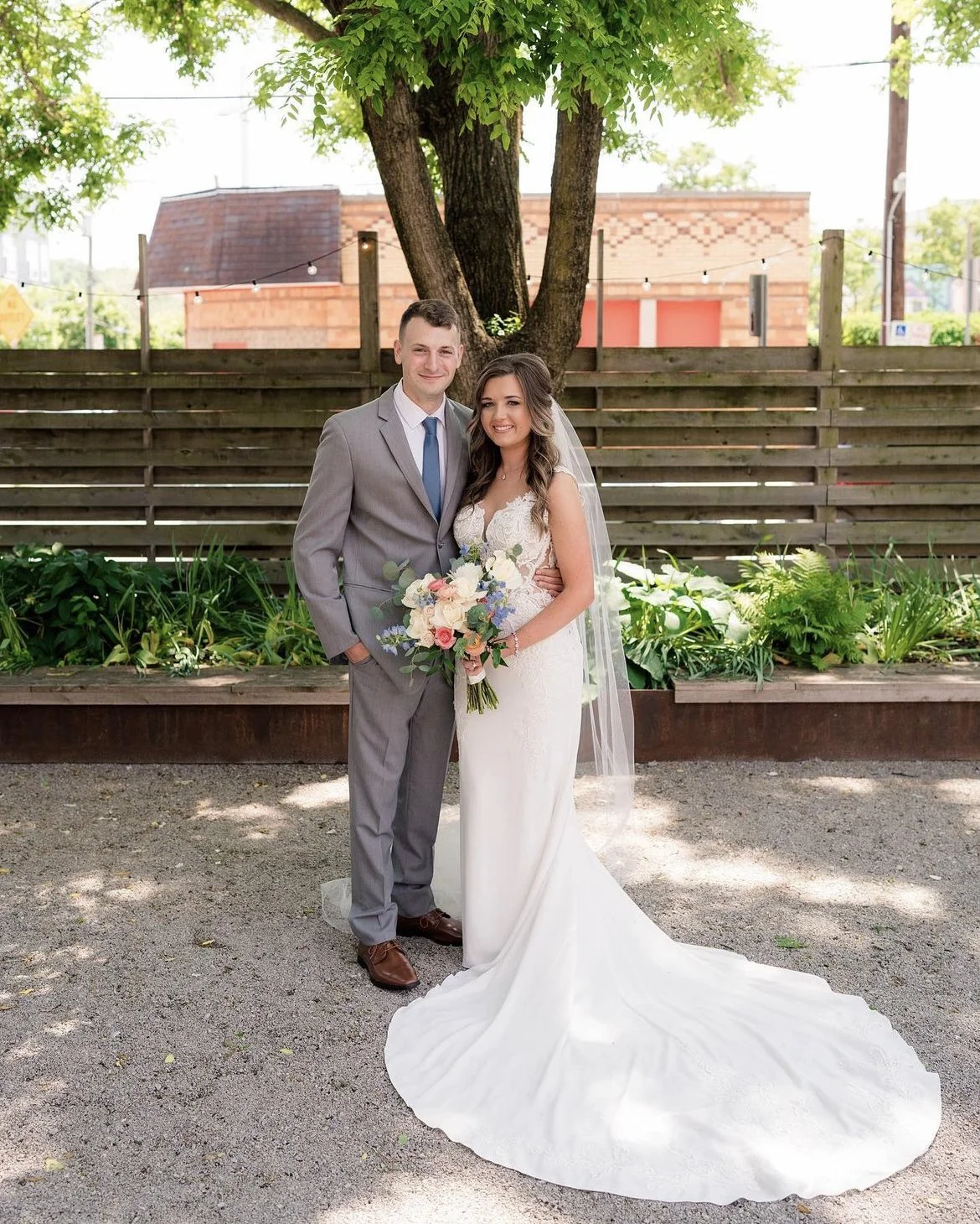 A bride and groom posing outdoors. The bride is holding a bouquet of flowers, and they stand under a large tree with a wooden fence in the background.